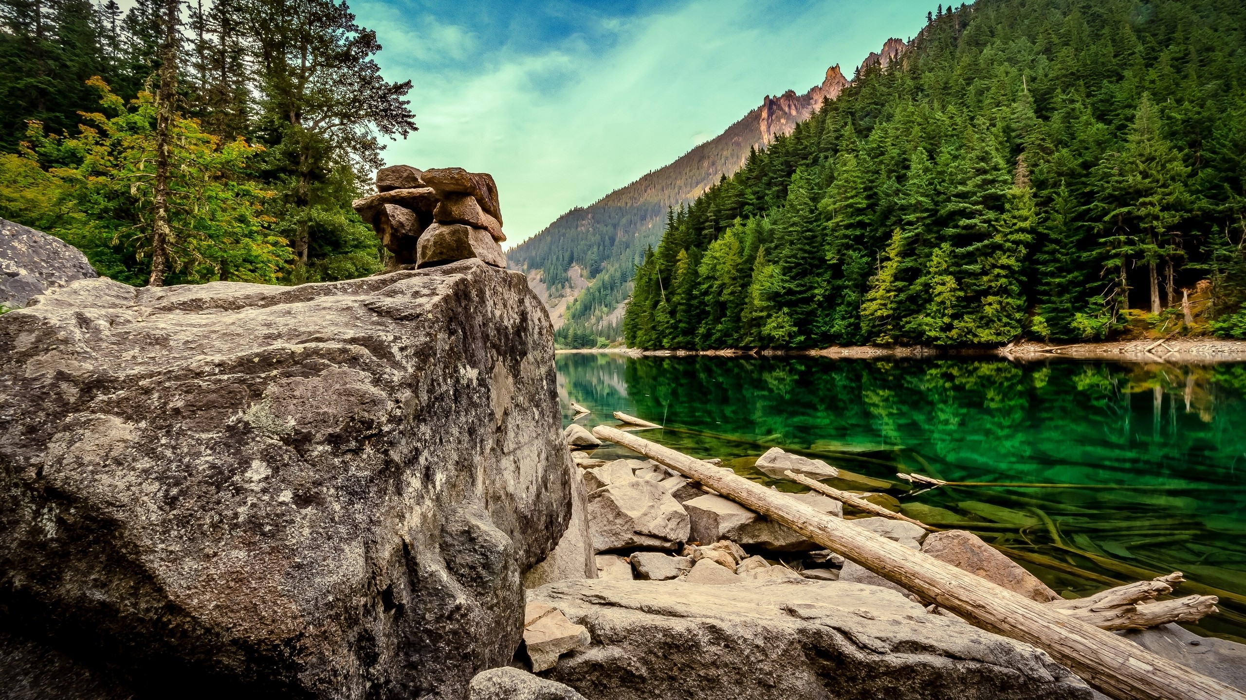 Rocky shore of a clear green lake with a forest and mountain backdrop HD Wallpaper