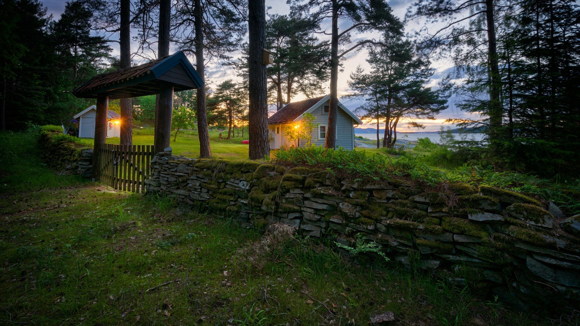 Stone wall and gate leading to houses by a lake at sunset HD Wallpaper