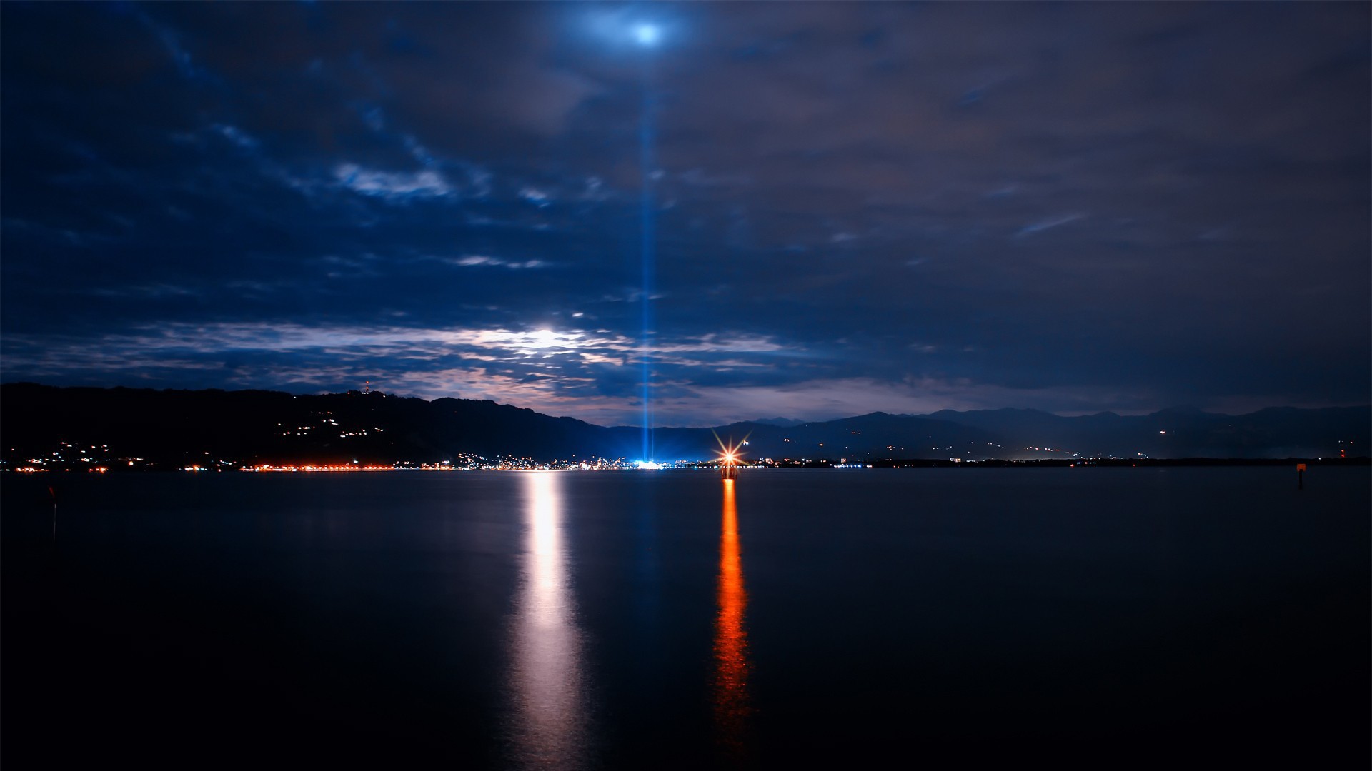 City lights reflected on a lake at night with a light beam HD Wallpaper