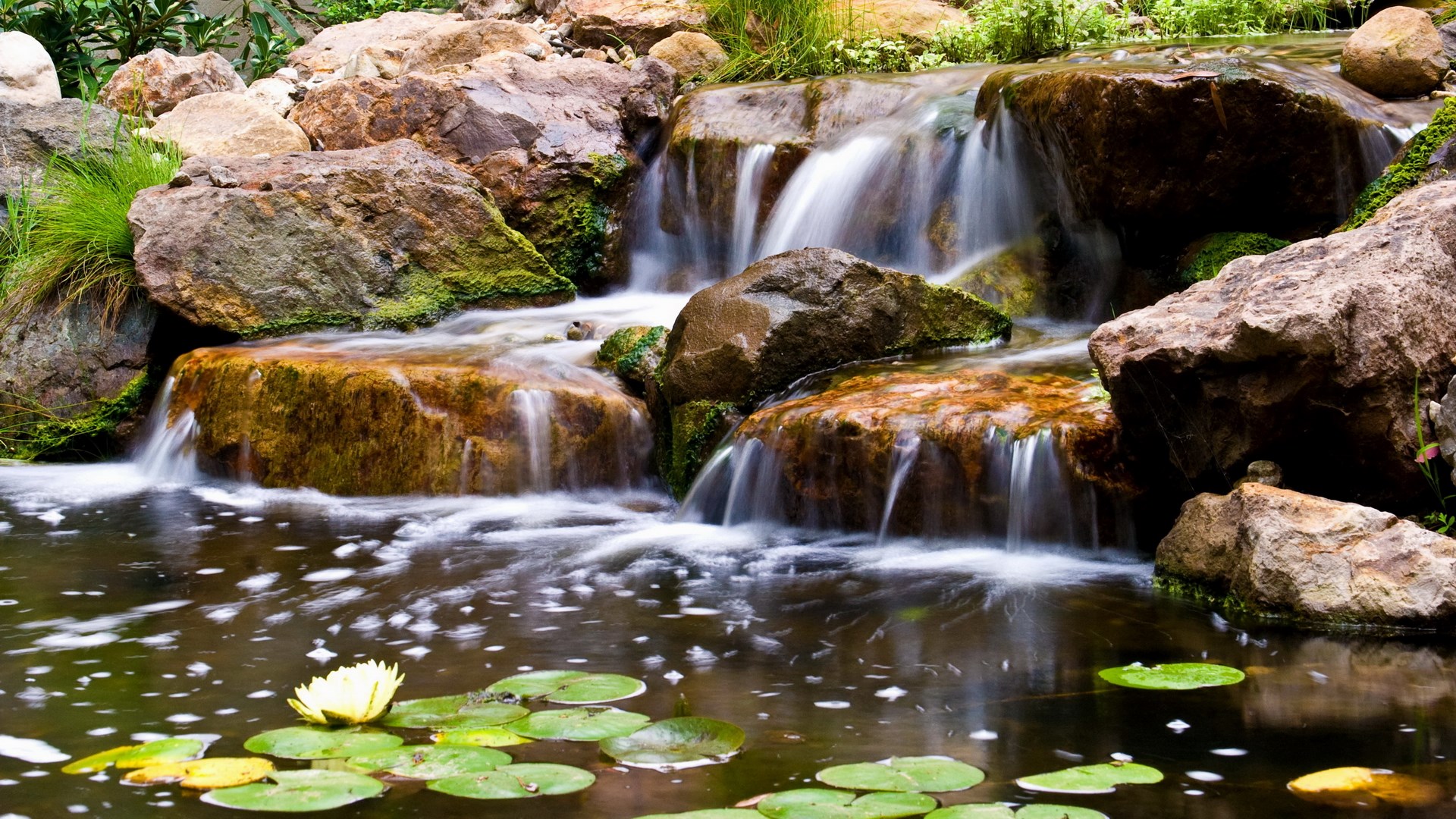 Waterfall cascading over rocks into a pond with water lilies HD Wallpaper