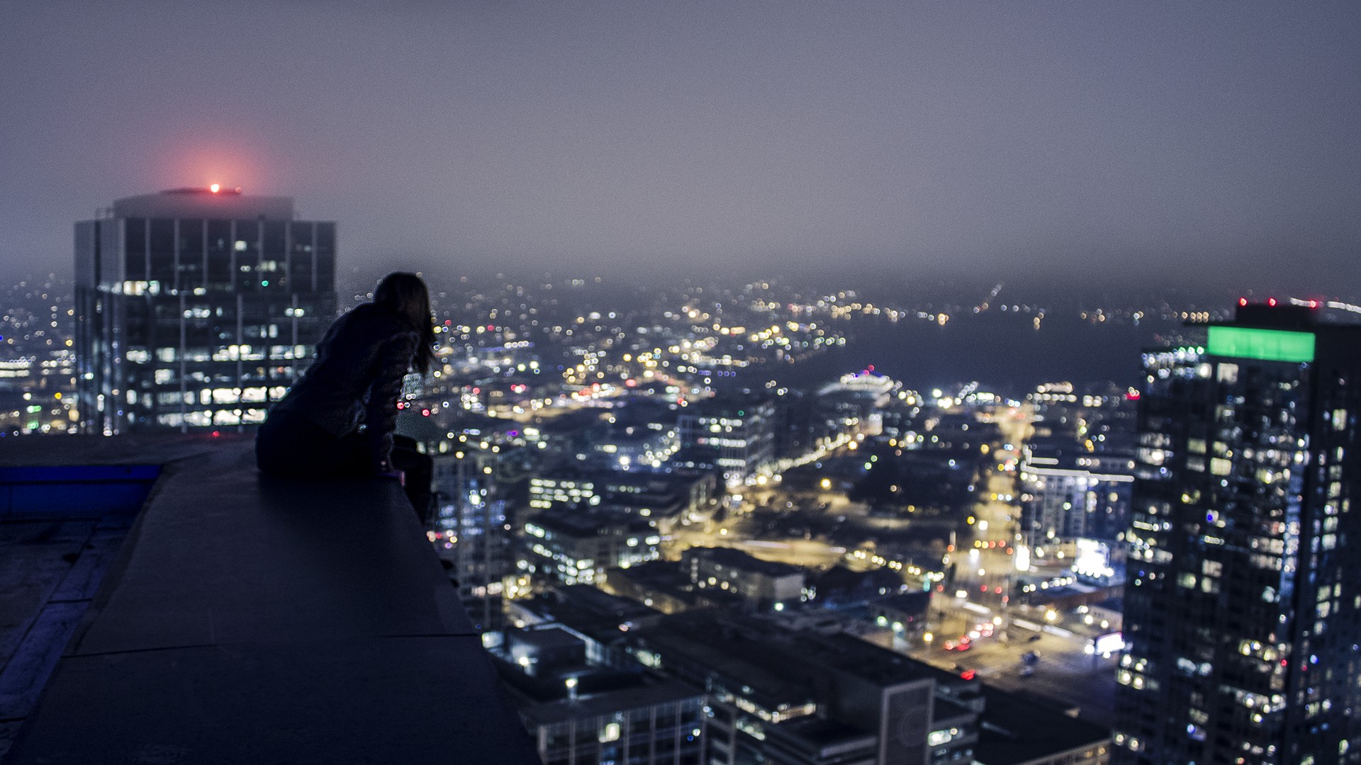 Person sitting on a rooftop overlooking a city at night HD Wallpaper