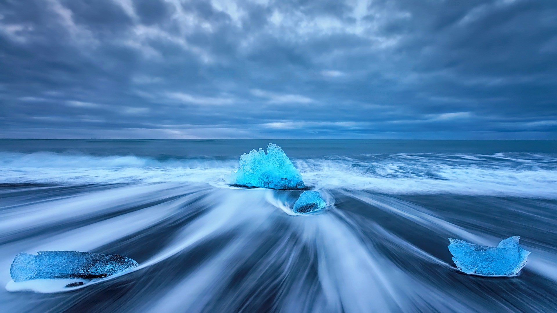 Blue icebergs on a black sand beach with waves HD Wallpaper
