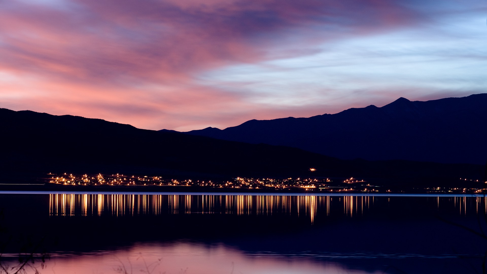 City lights reflected on a calm lake at sunset with mountains HD Wallpaper