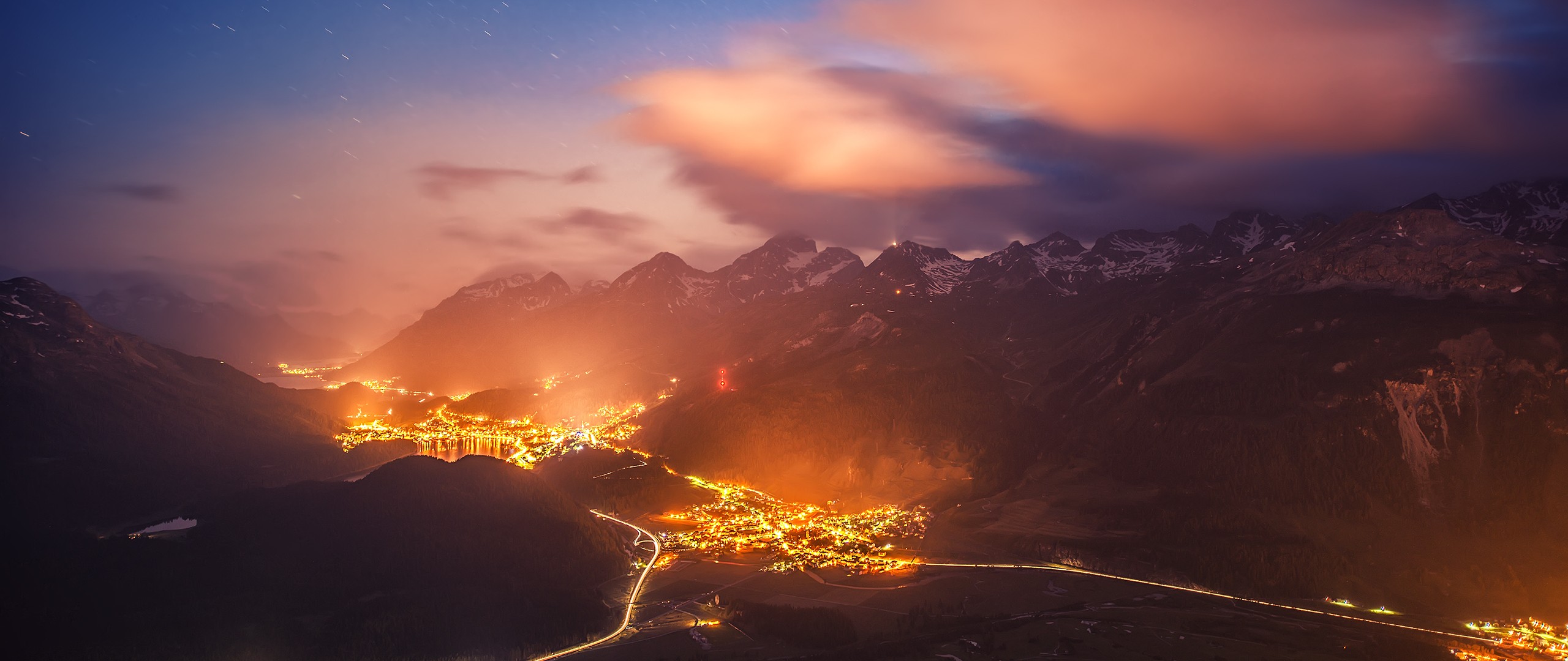 Town lights illuminate valley below snow-capped mountains at night HD Wallpaper