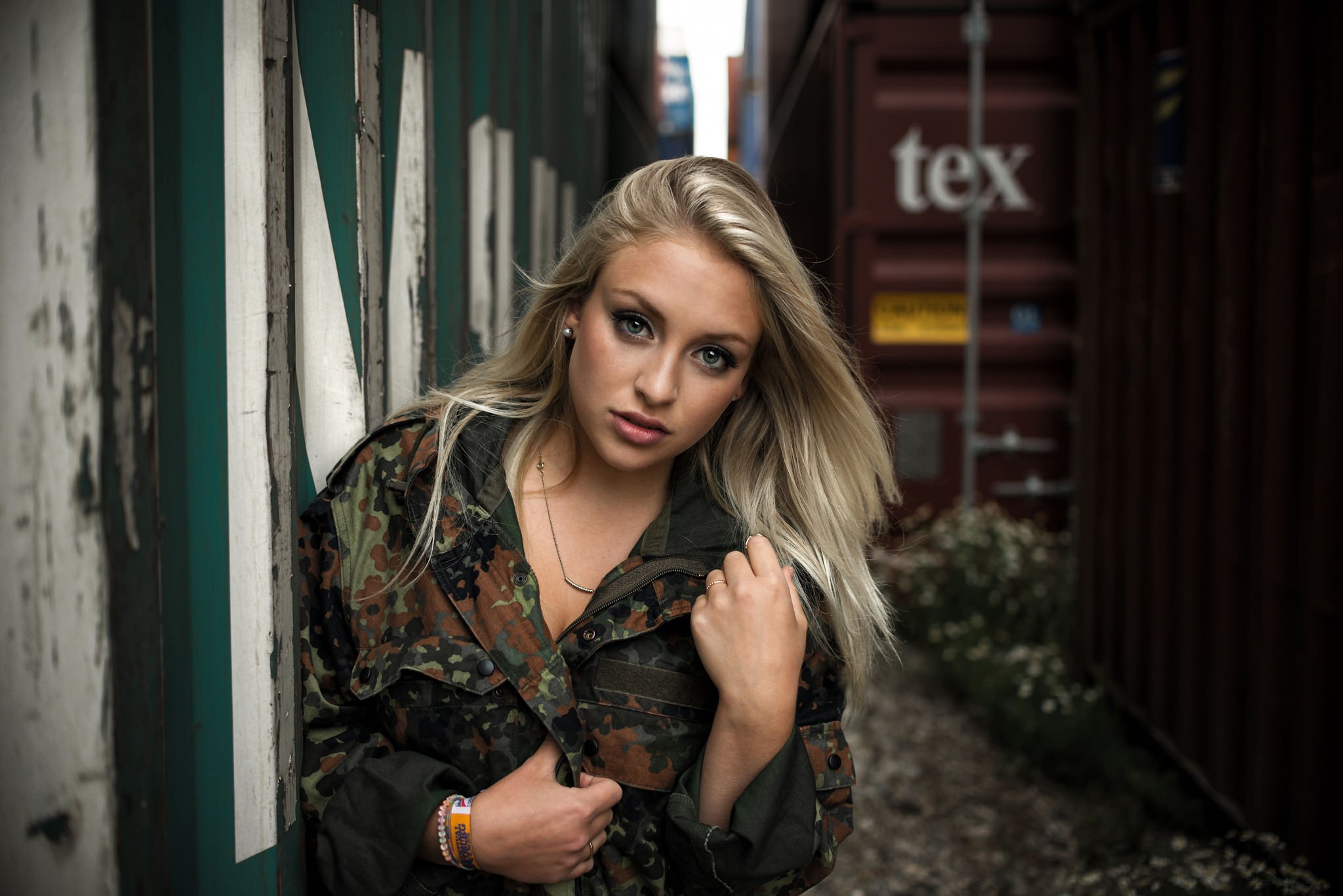 Woman in camouflage jacket leaning against shipping containers HD Wallpaper