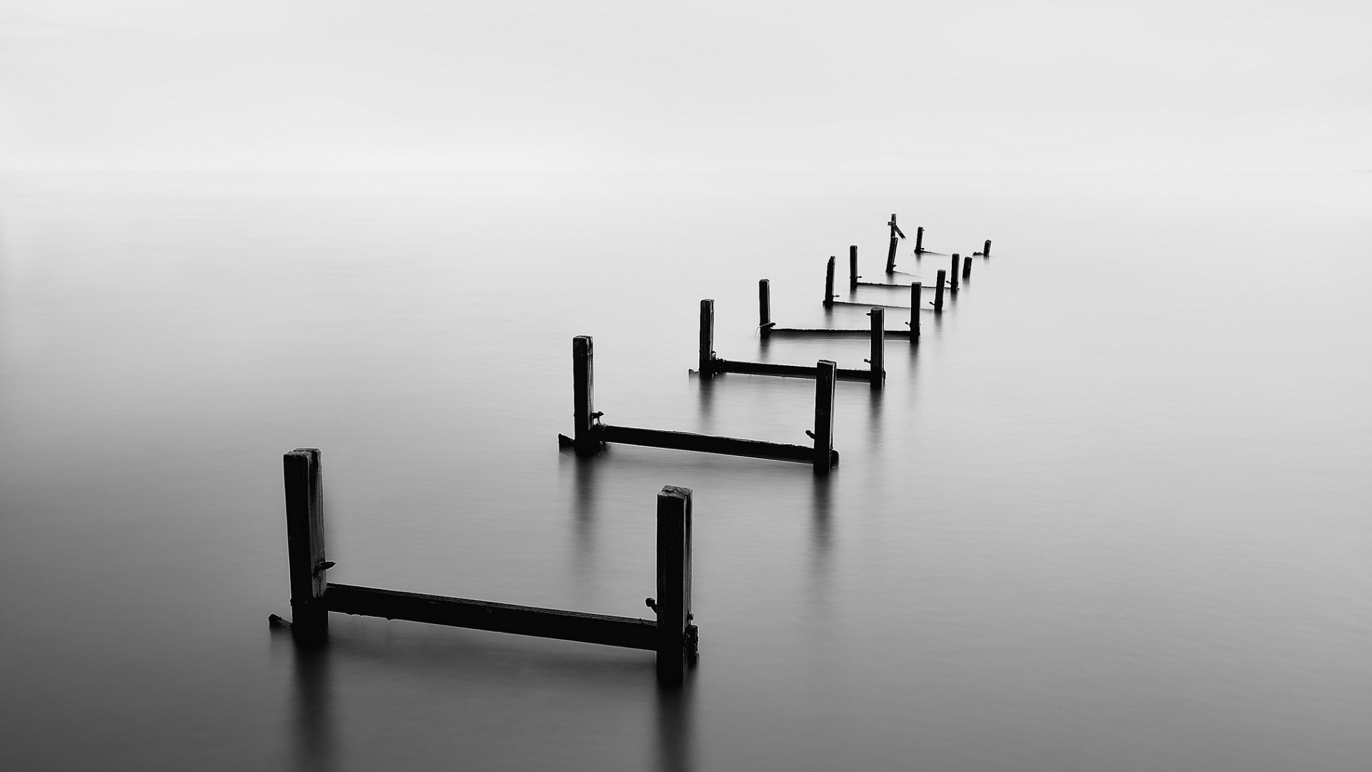 Monochrome long exposure of wooden pier ruins in calm water HD Wallpaper