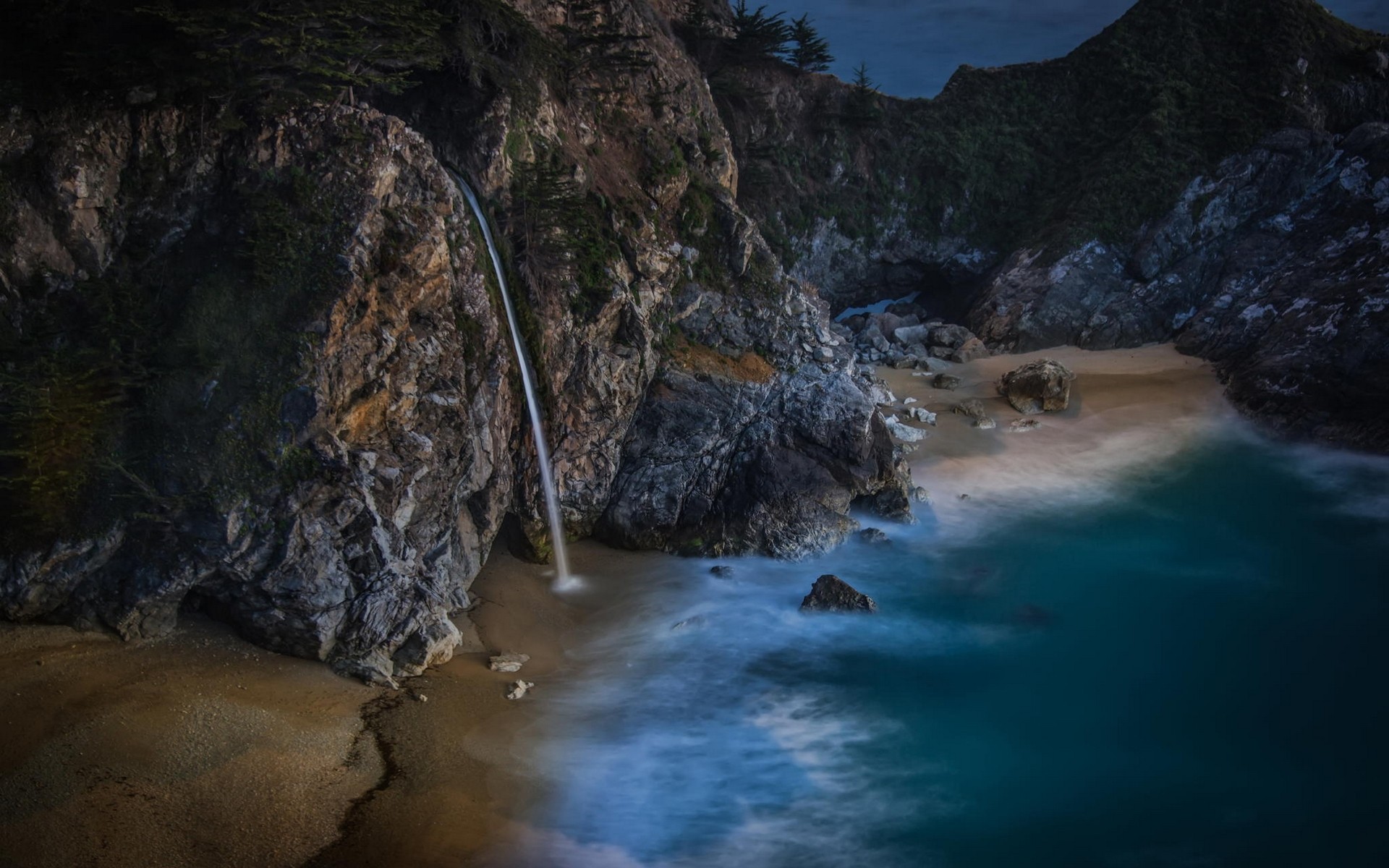 Waterfall cascading onto a sandy beach by the sea HD Wallpaper