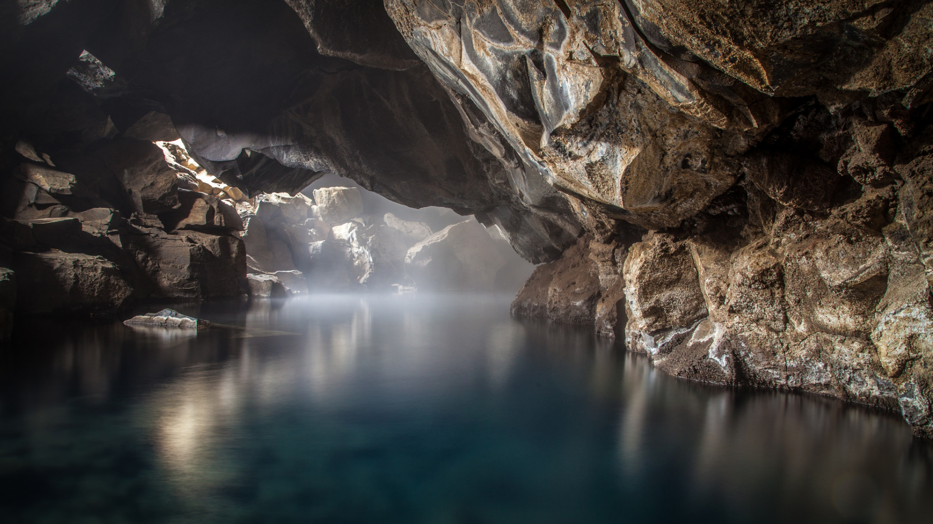Sunlight filtering into a misty cave with a dark blue lake HD Wallpaper