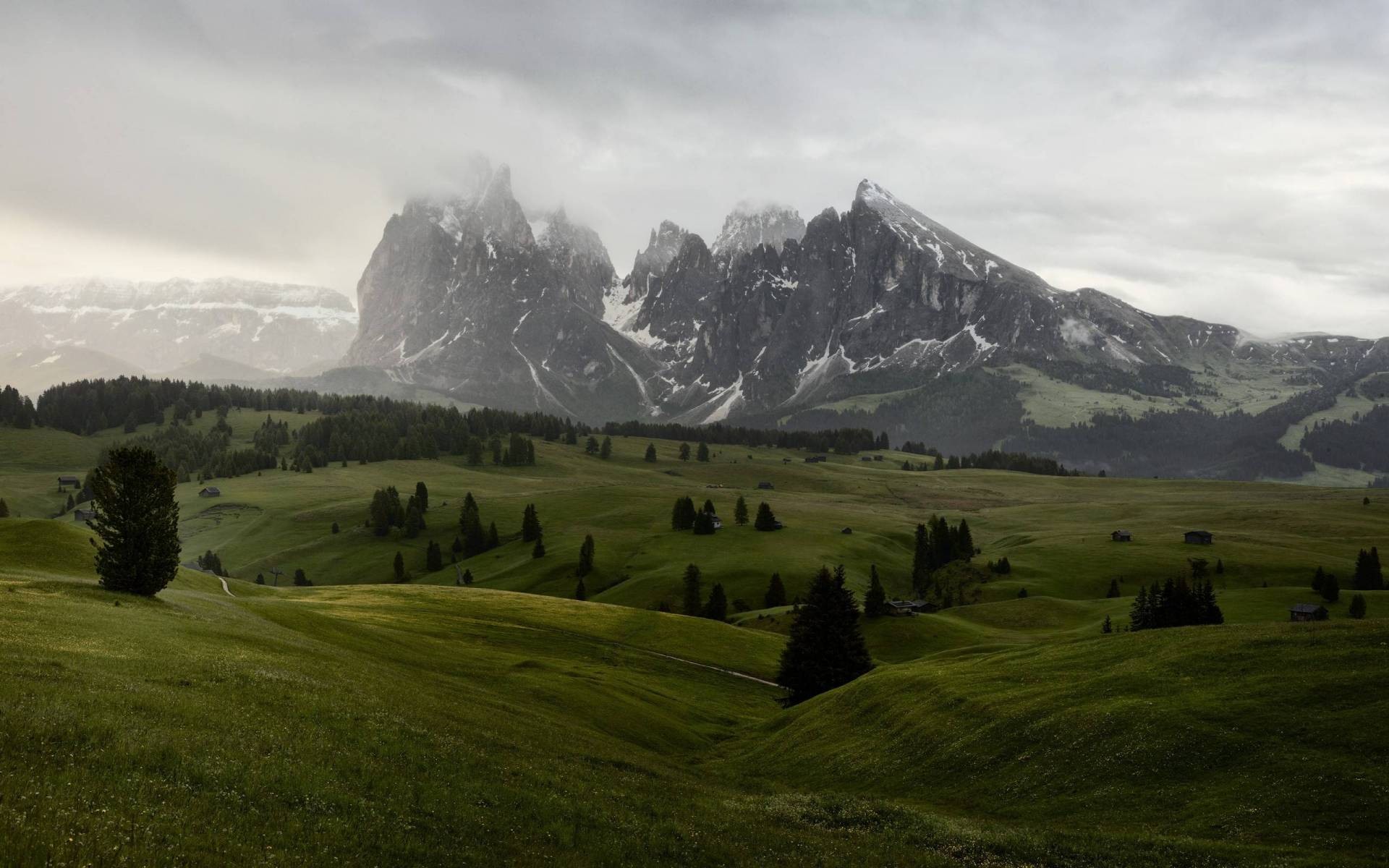 Rolling green hills and jagged mountains under a cloudy sky HD Wallpaper