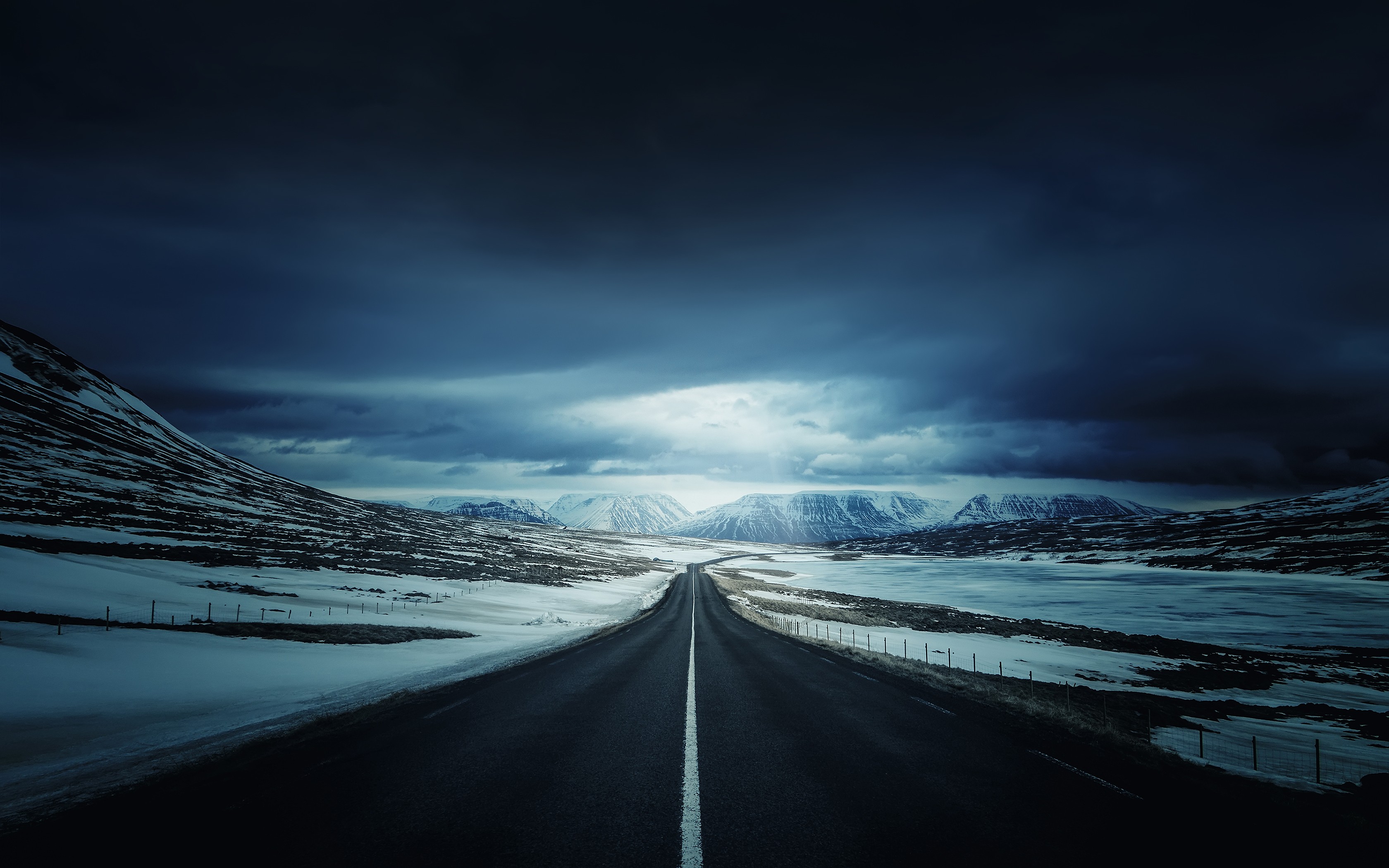 A lonely road through a snowy, mountainous landscape under a dark sky. HD Wallpaper
