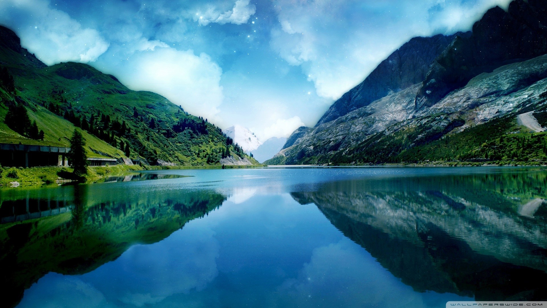 A serene lake reflects majestic mountains under a cloudy sky. HD Wallpaper