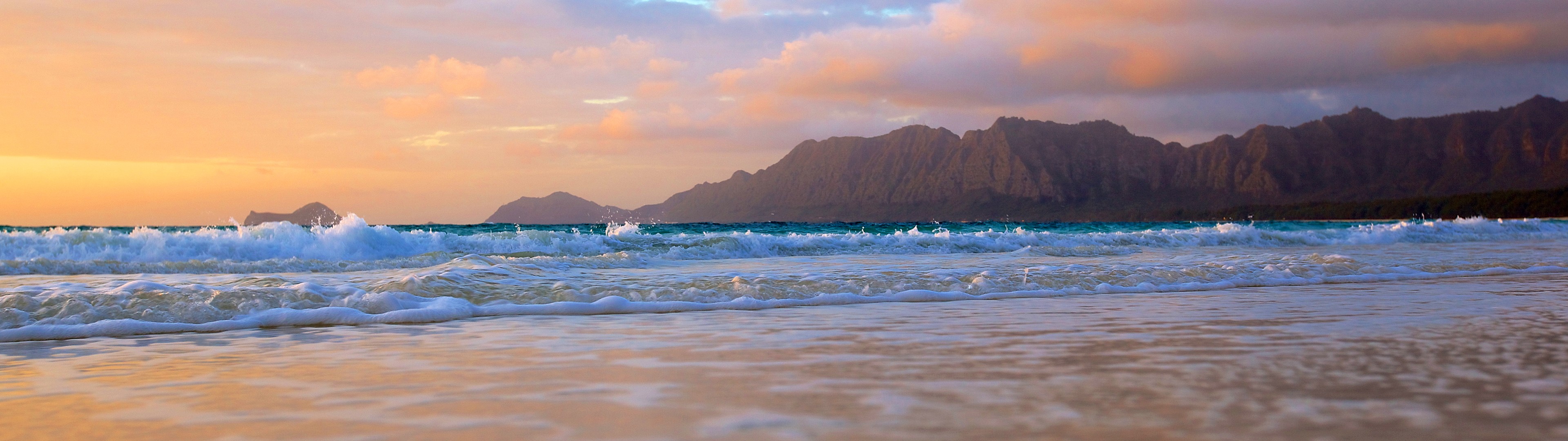 Ocean waves crash on a beach with mountains in the background at sunset. HD Wallpaper