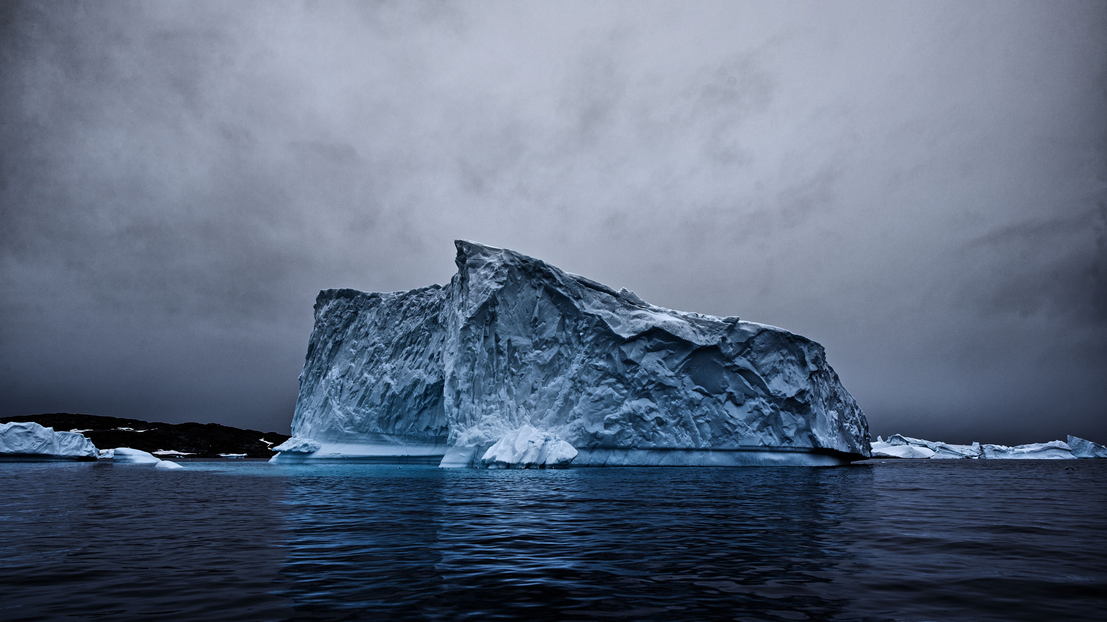 Large iceberg floating in dark water under cloudy sky HD Wallpaper