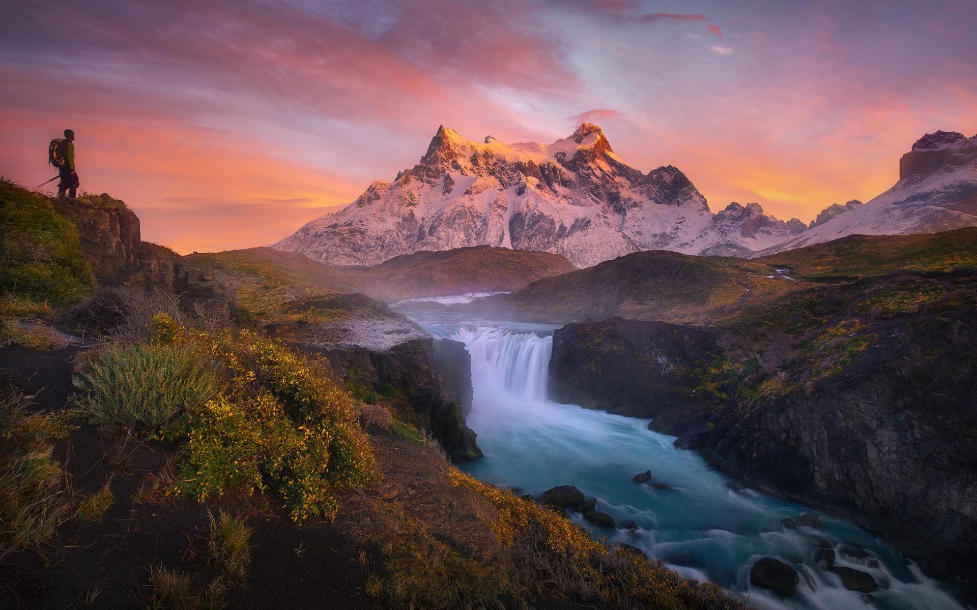 Hiker overlooks waterfall and snowy mountains at sunrise HD Wallpaper