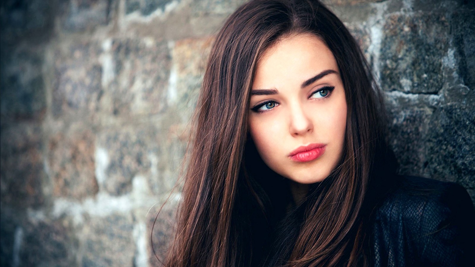 Brunette woman with blue eyes leaning against a stone wall HD Wallpaper