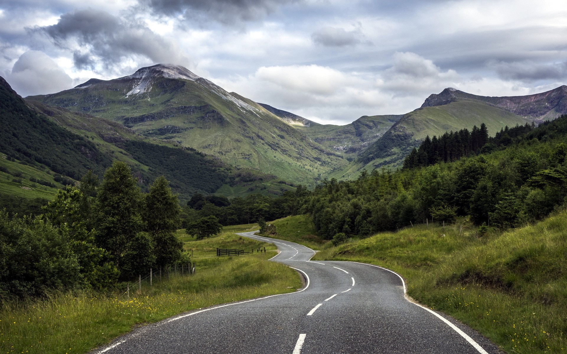 Winding road through mountains and forest under cloudy sky HD Wallpaper