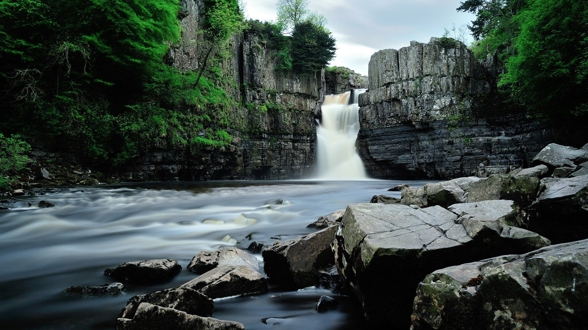 Waterfall cascading over rocky cliffs into a river HD Wallpaper