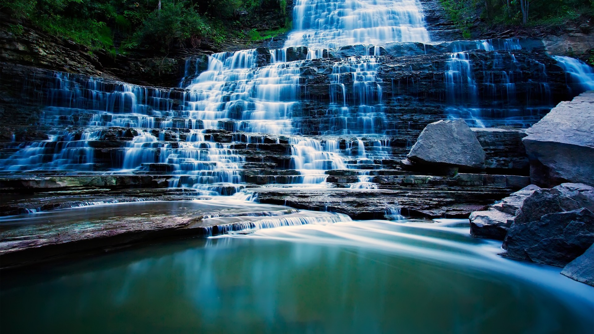 A cascading waterfall flows over layered rocks into a pool HD Wallpaper