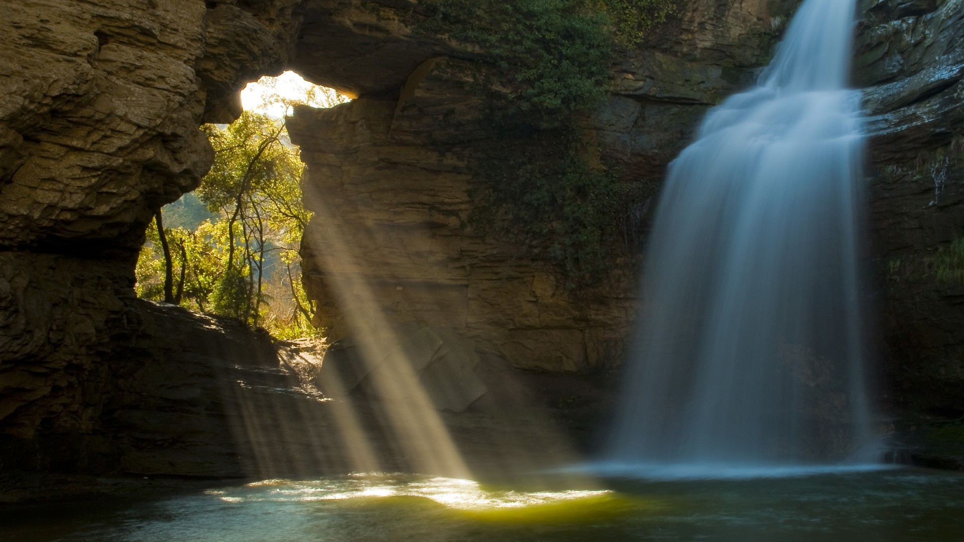 Waterfall with sun rays through rock arch HD Wallpaper