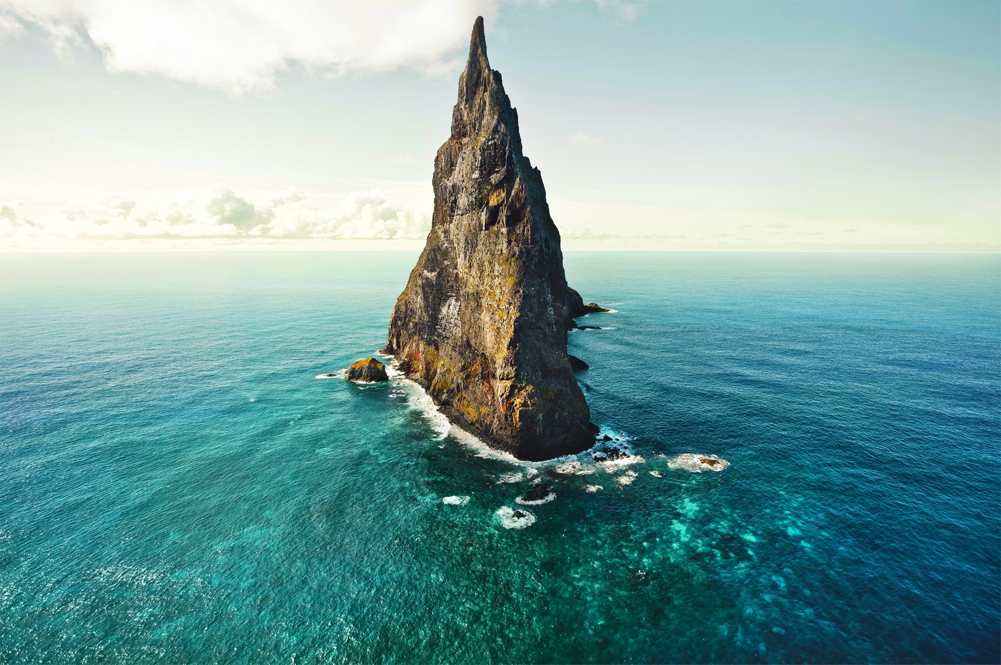 A tall, jagged rock formation rises from the sea. HD Wallpaper