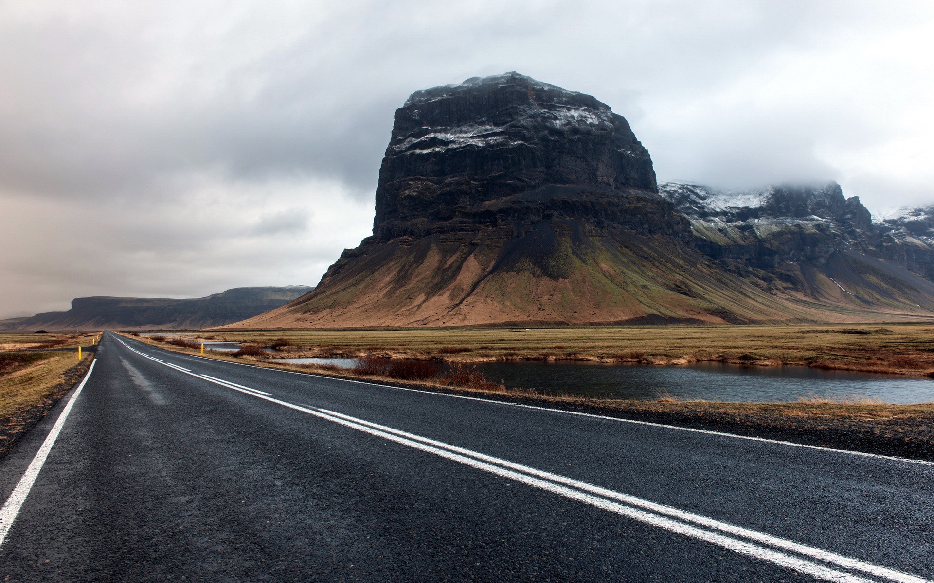 Asphalt road leading to a large mountain under a cloudy sky HD Wallpaper