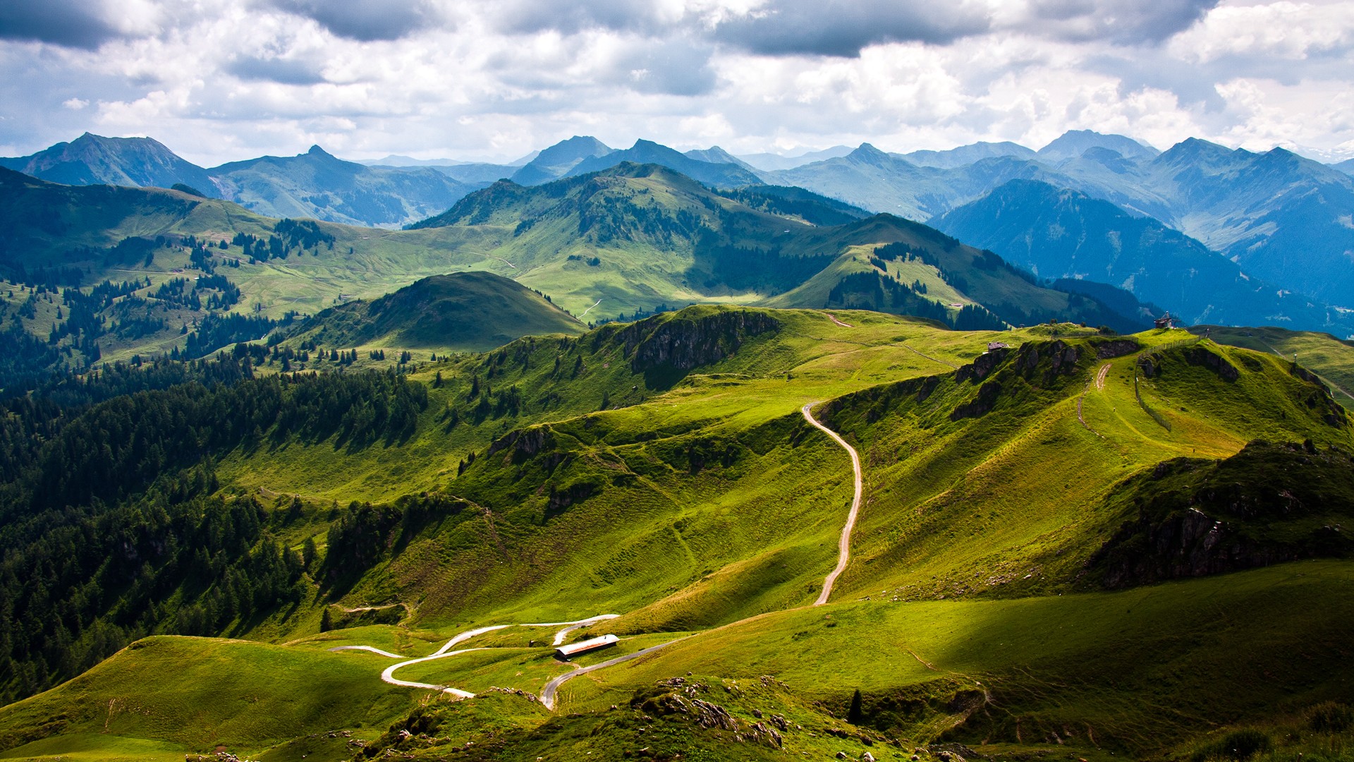 Green mountains with winding paths and a cloudy sky HD Wallpaper