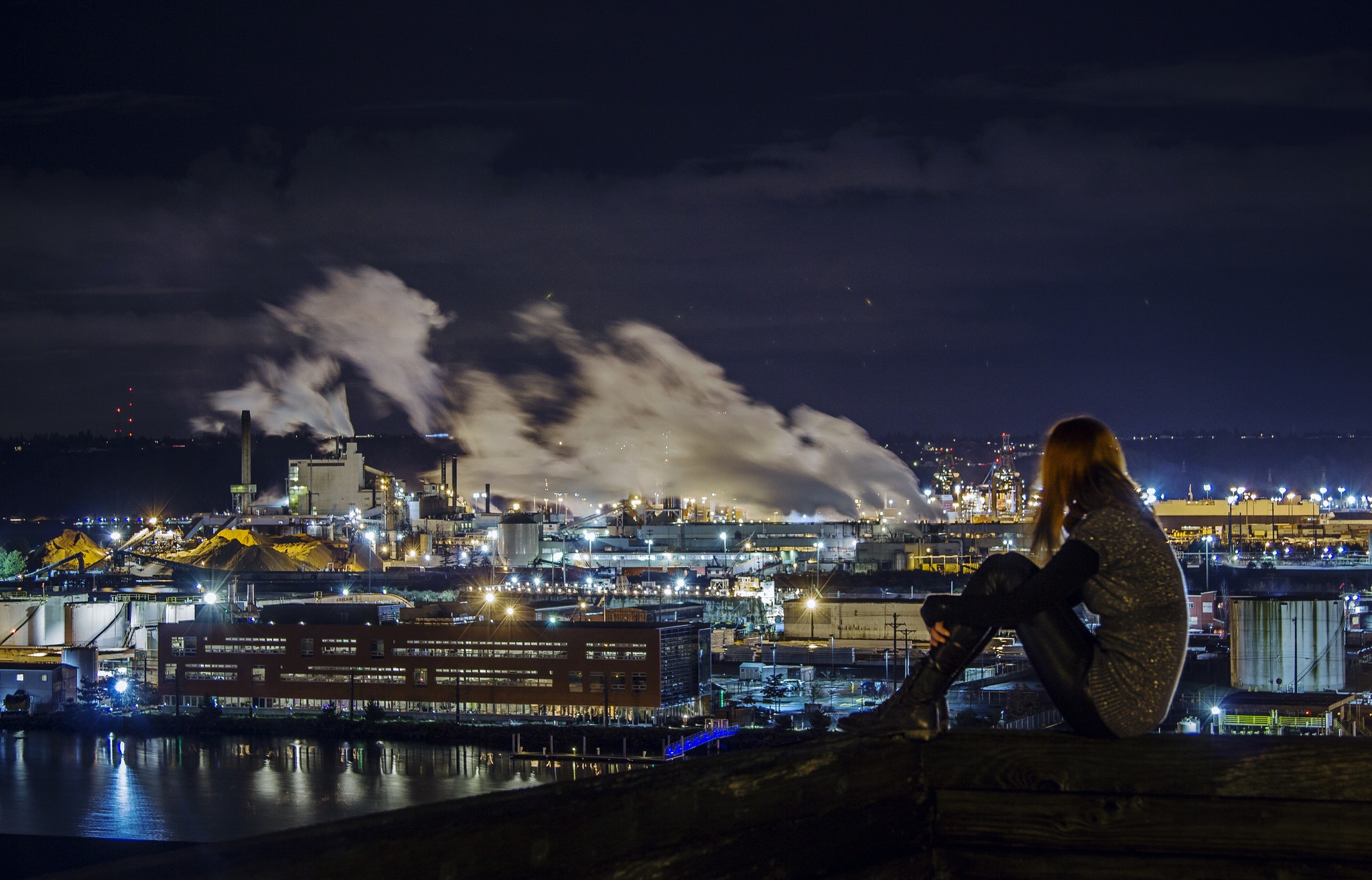 Woman sitting on a ledge overlooking an industrial cityscape at night HD Wallpaper