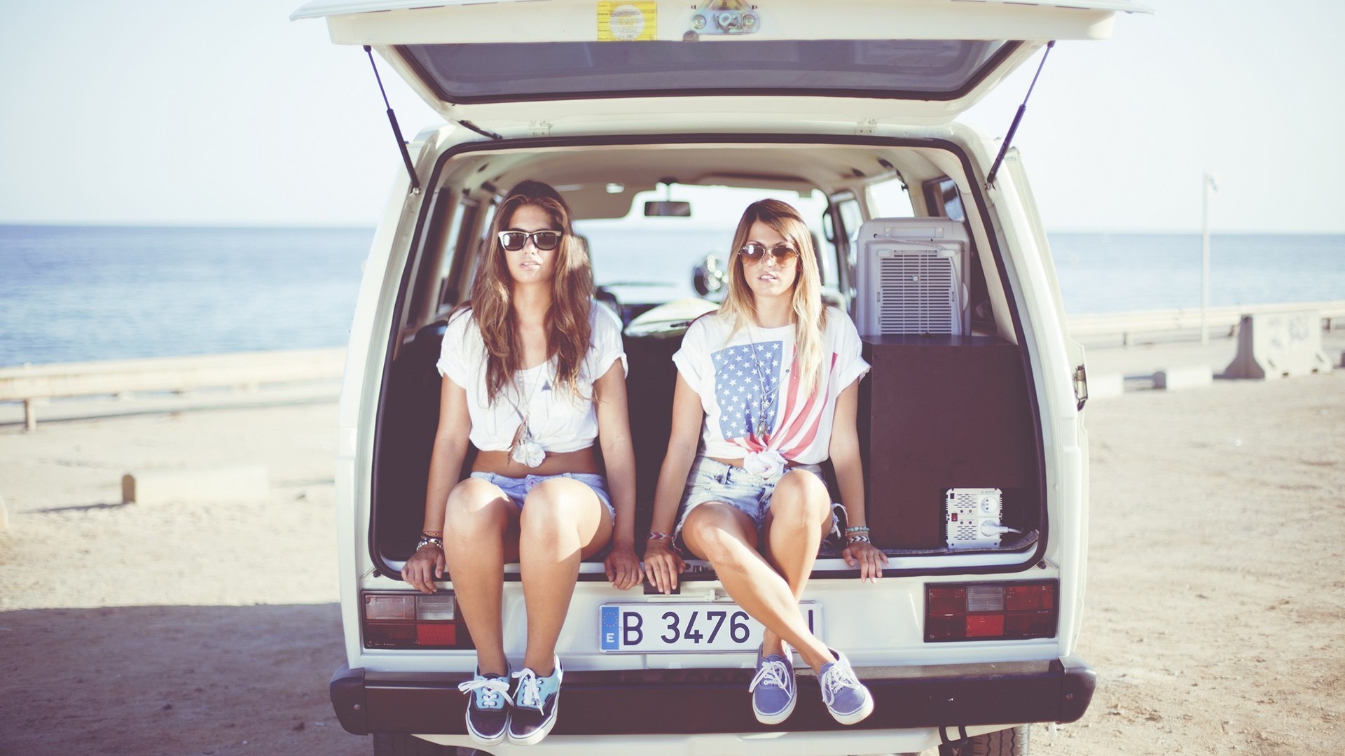 Two women in jean shorts sitting in the back of a Volkswagen van at the beach HD Wallpaper