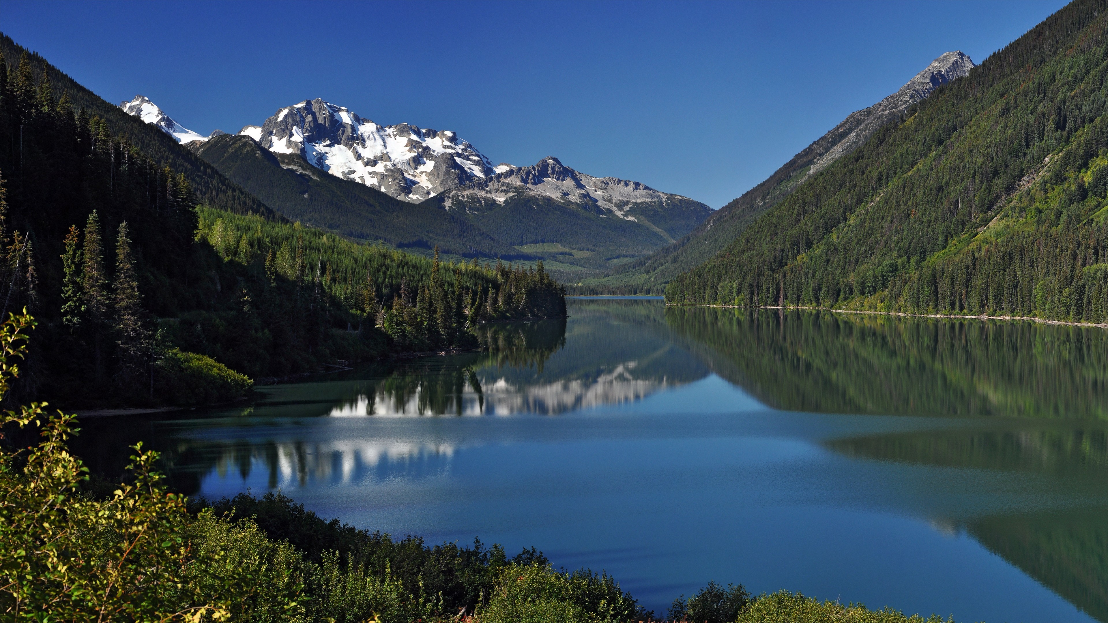 Snow-capped mountains reflected in a calm lake surrounded by forests HD Wallpaper