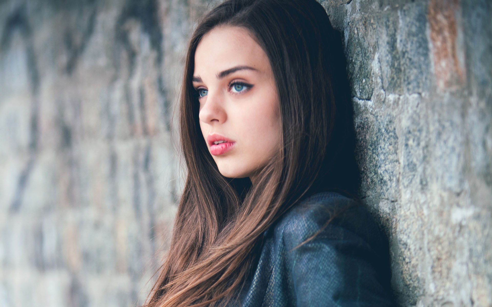 Young woman with blue eyes leaning against a stone wall HD Wallpaper