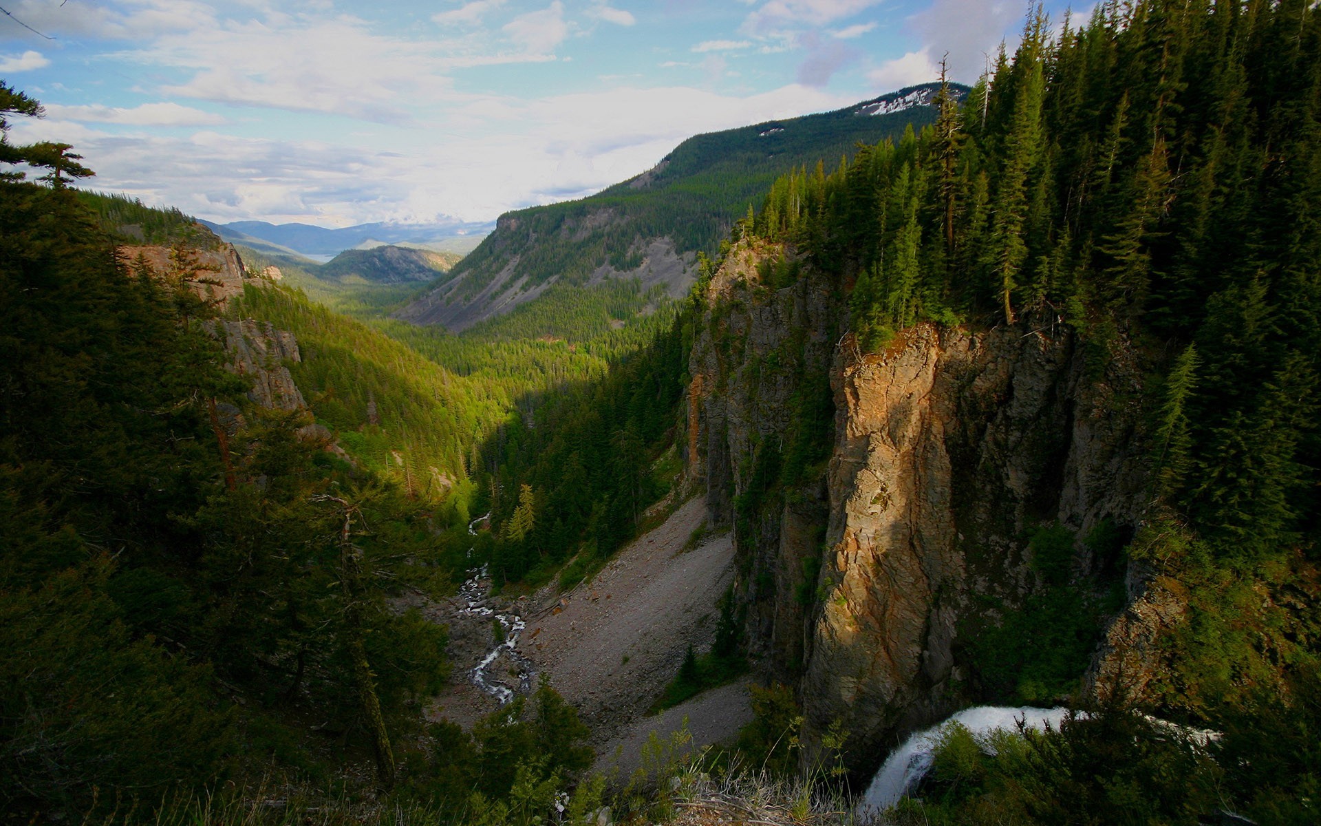 A waterfall cascades over a cliff in a forested mountain valley. HD Wallpaper
