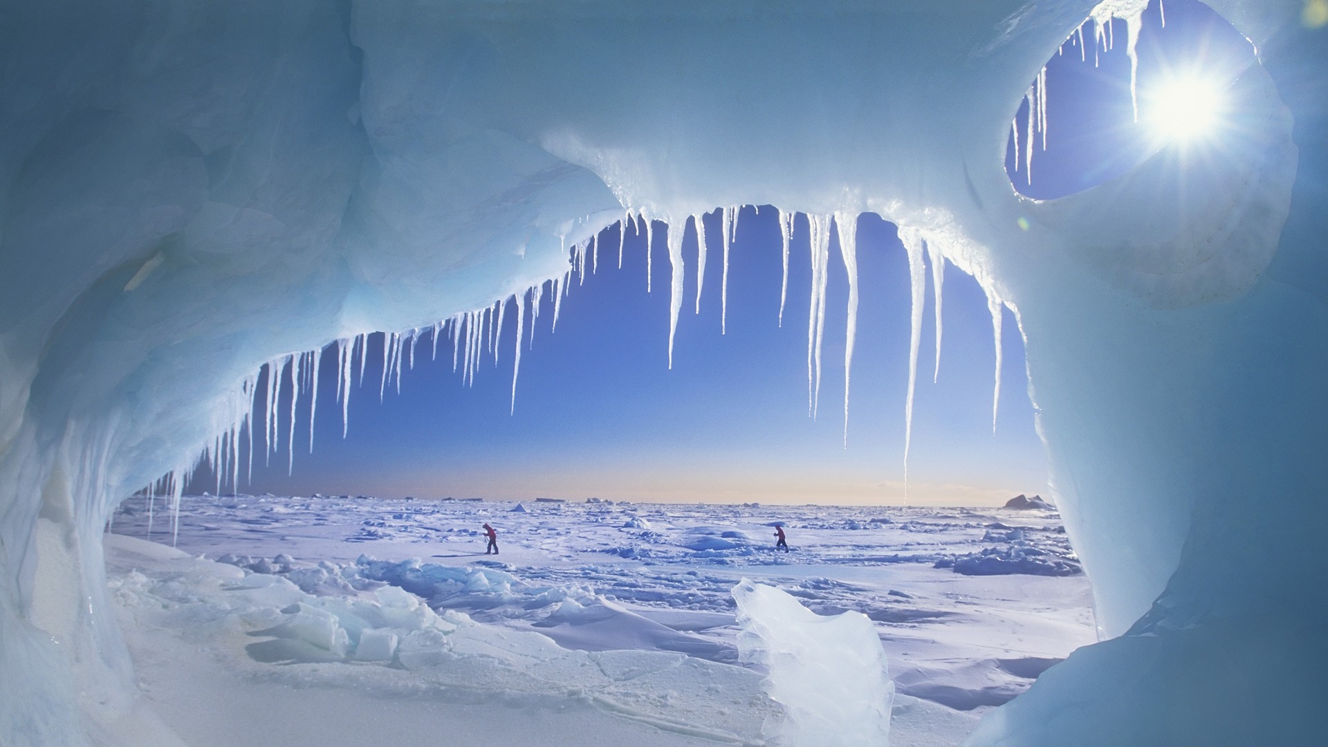 Two people walk across an icy landscape under a bright sun. HD Wallpaper