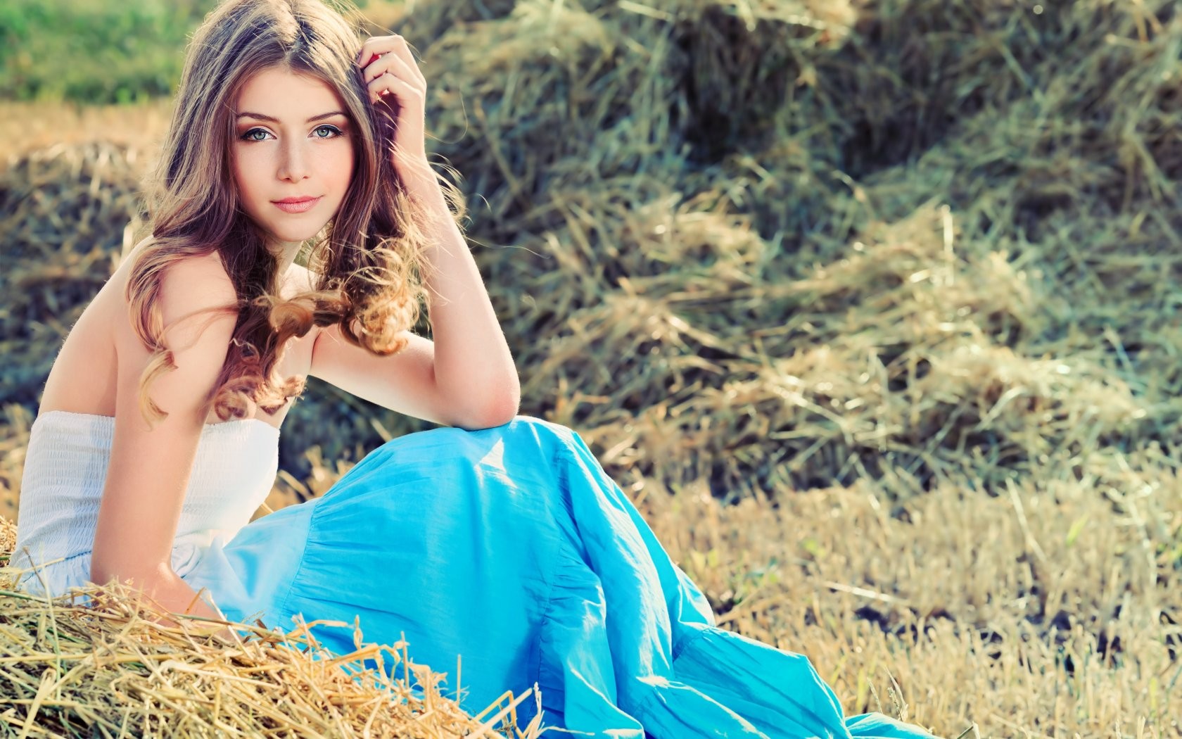 Young woman with long brown hair in a blue dress sitting in hay HD Wallpaper