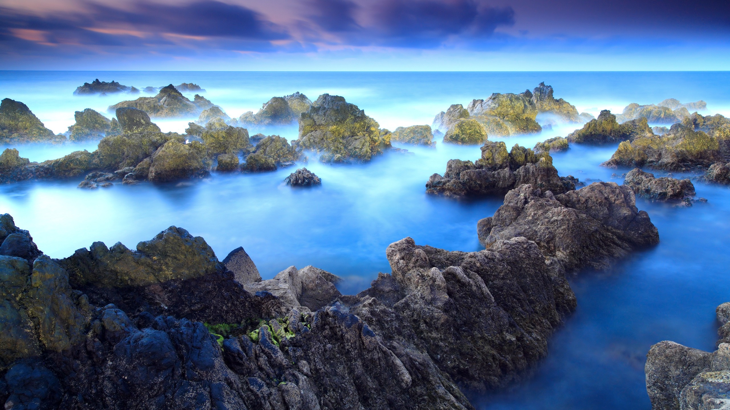Rocky coastline with misty blue ocean and purple clouds HD Wallpaper