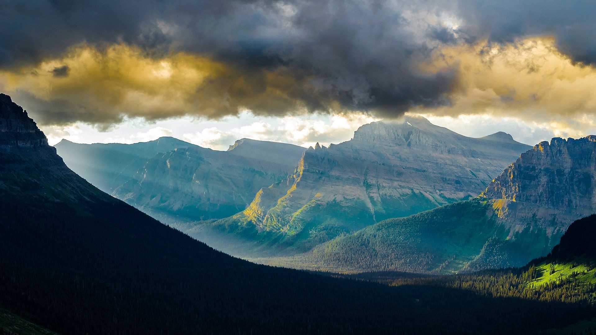 Sunlight breaking through clouds over mountains in Glacier National Park HD Wallpaper