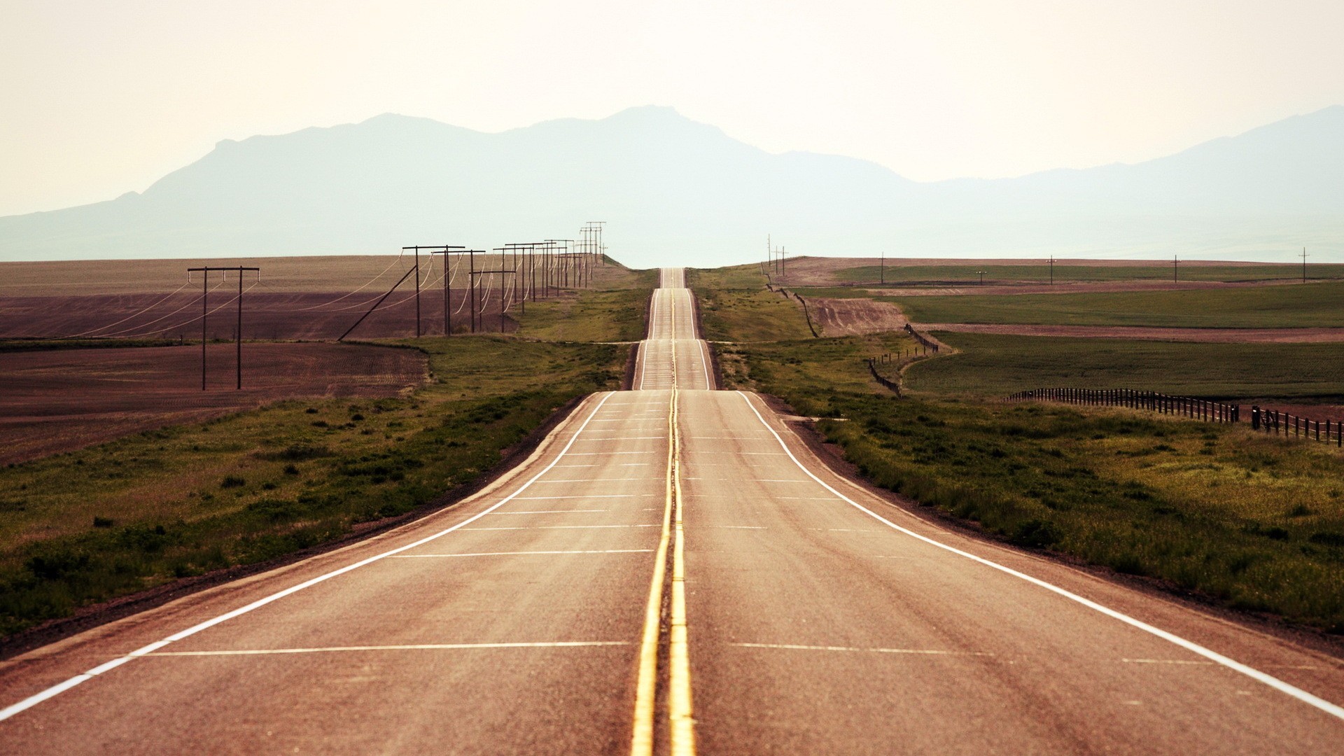 A long, empty highway stretches towards distant mountains. HD Wallpaper