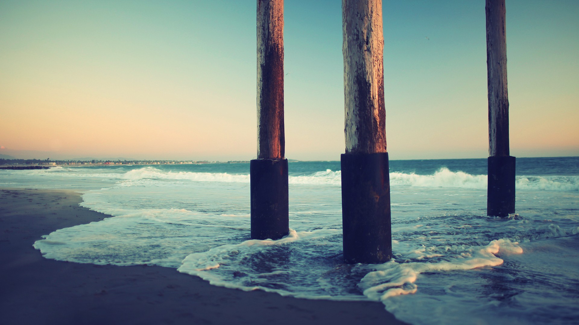Ocean waves crash against pier pilings on a beach HD Wallpaper