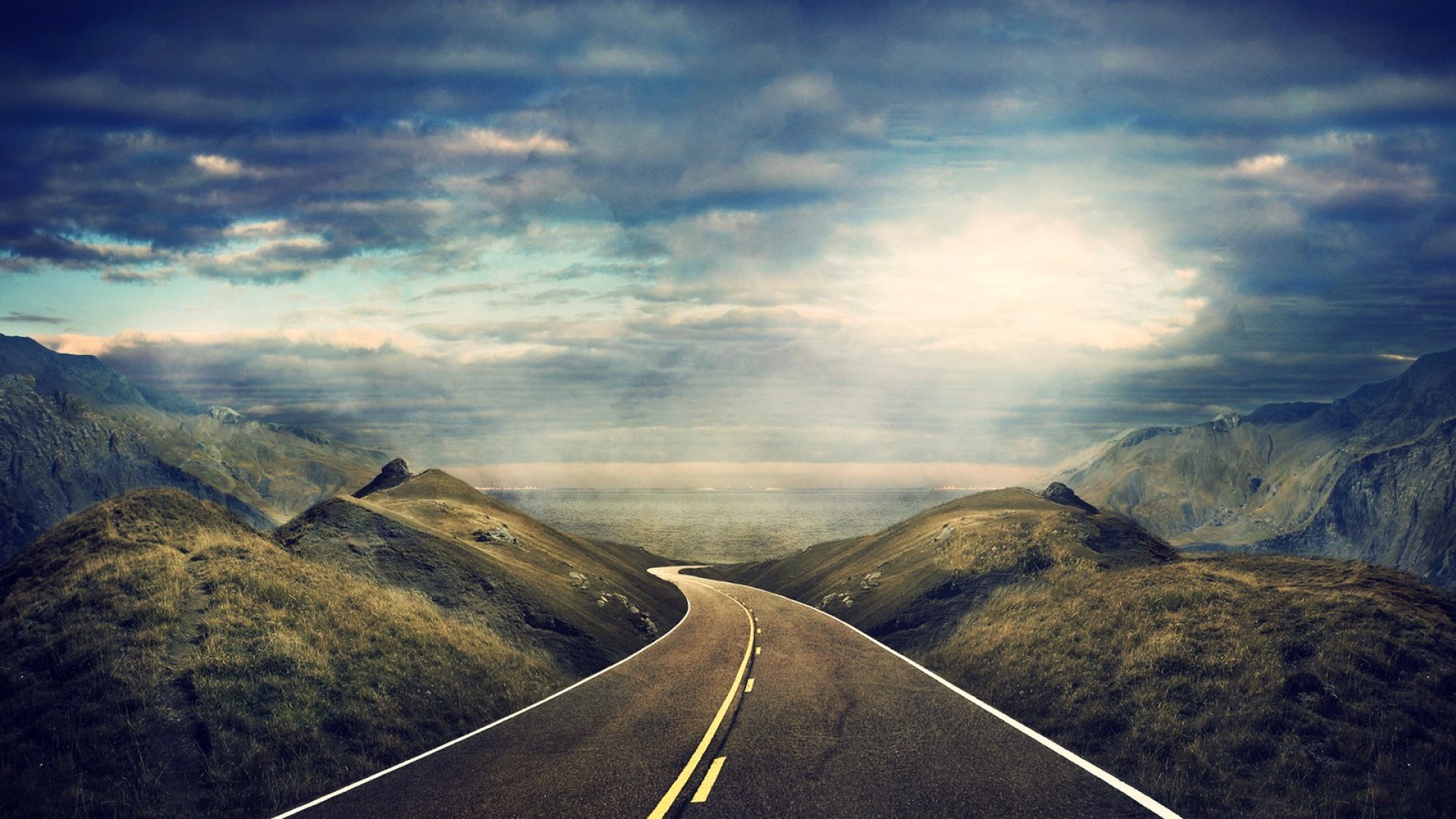 A winding road through mountainous landscape under a dramatic sky HD Wallpaper