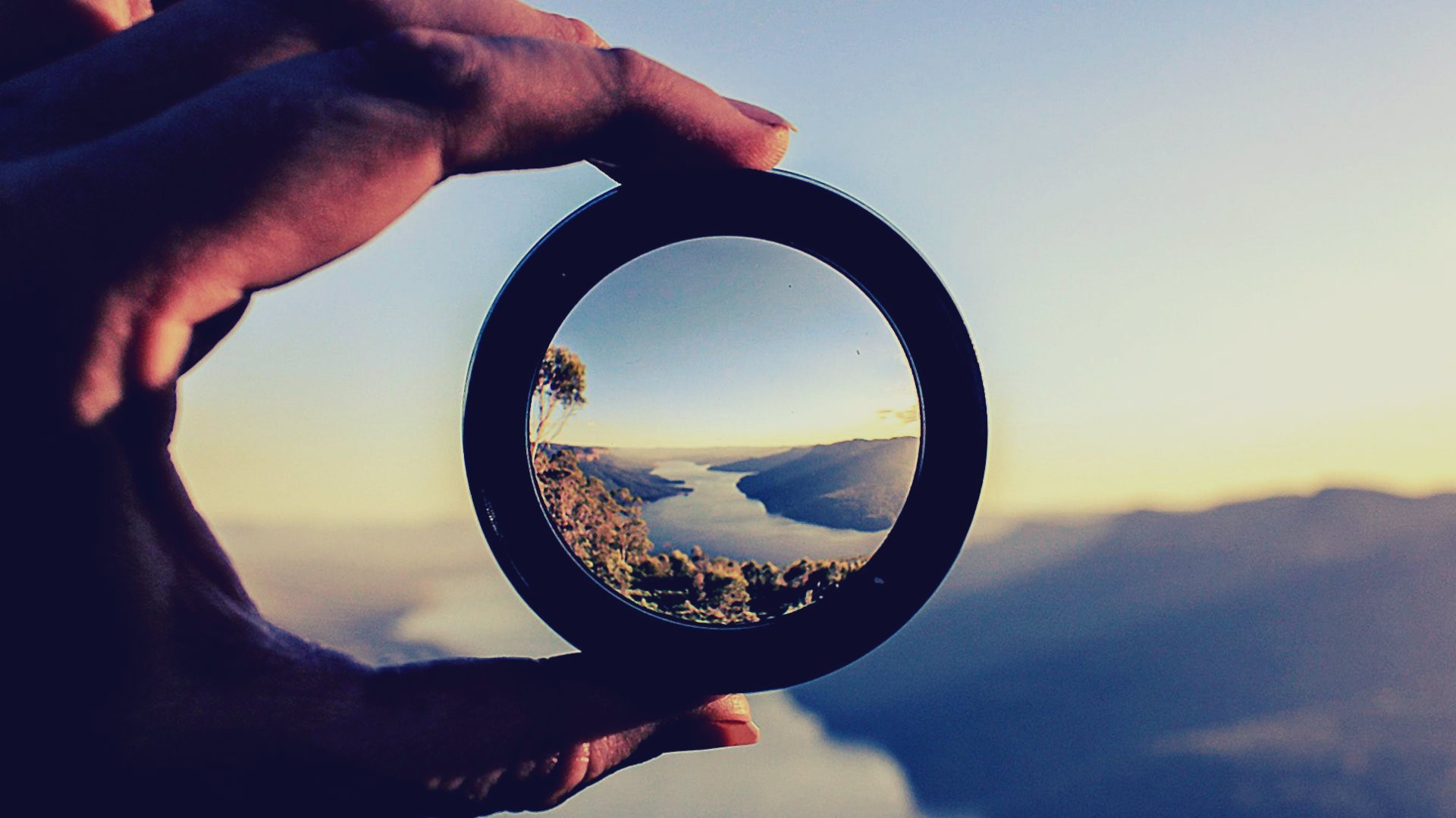 Hand holding a lens framing a river landscape HD Wallpaper