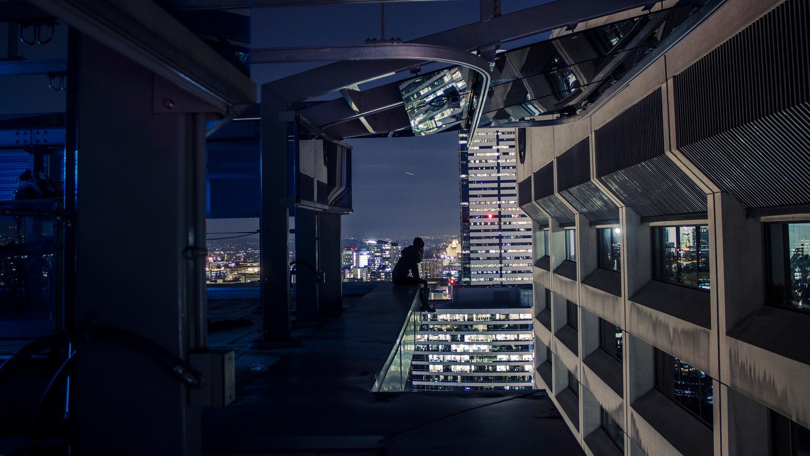 Person sitting on a skyscraper rooftop at night overlooking a cityscape HD Wallpaper