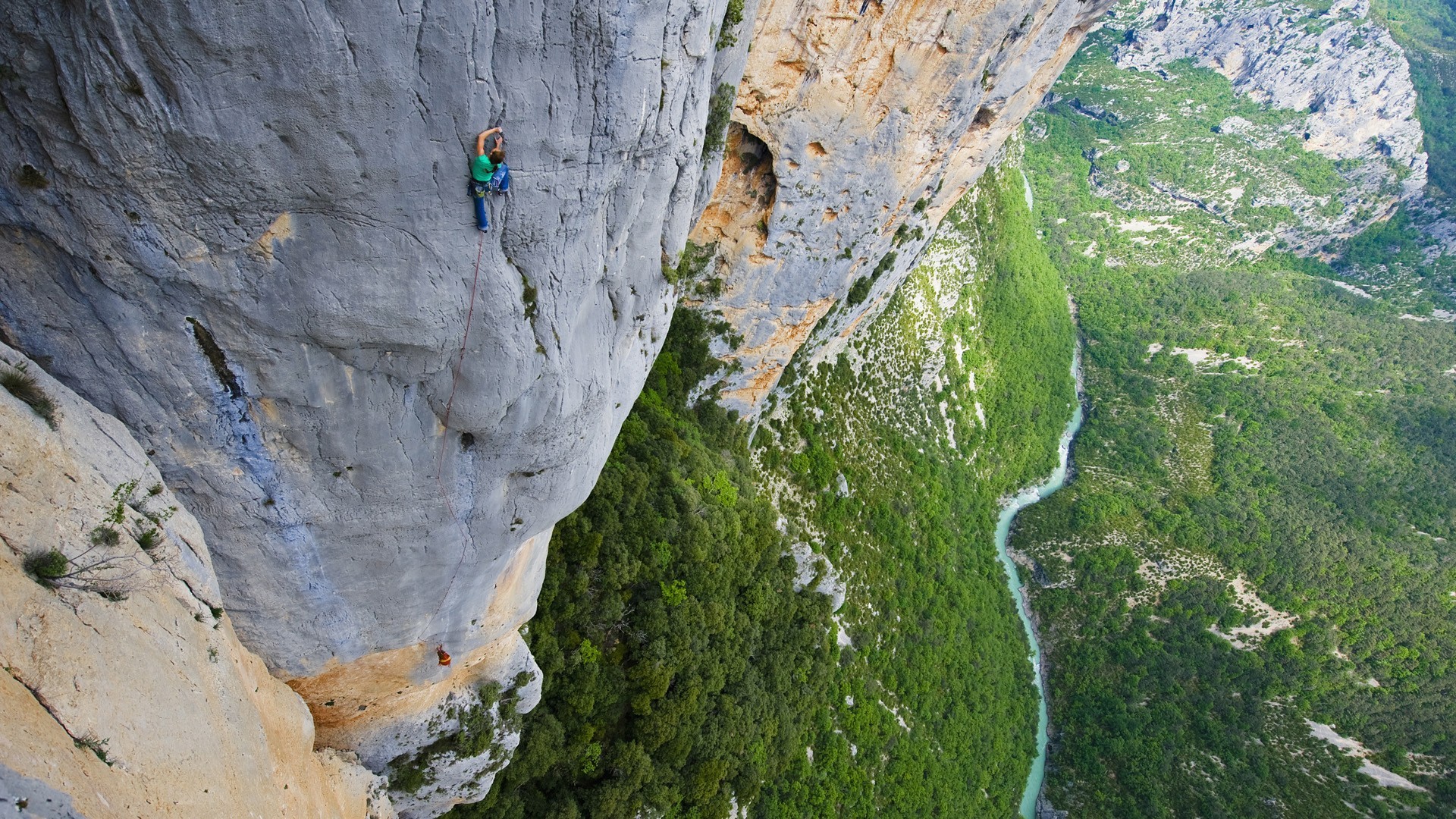Rock climber on a sheer cliff face above a green canyon HD Wallpaper