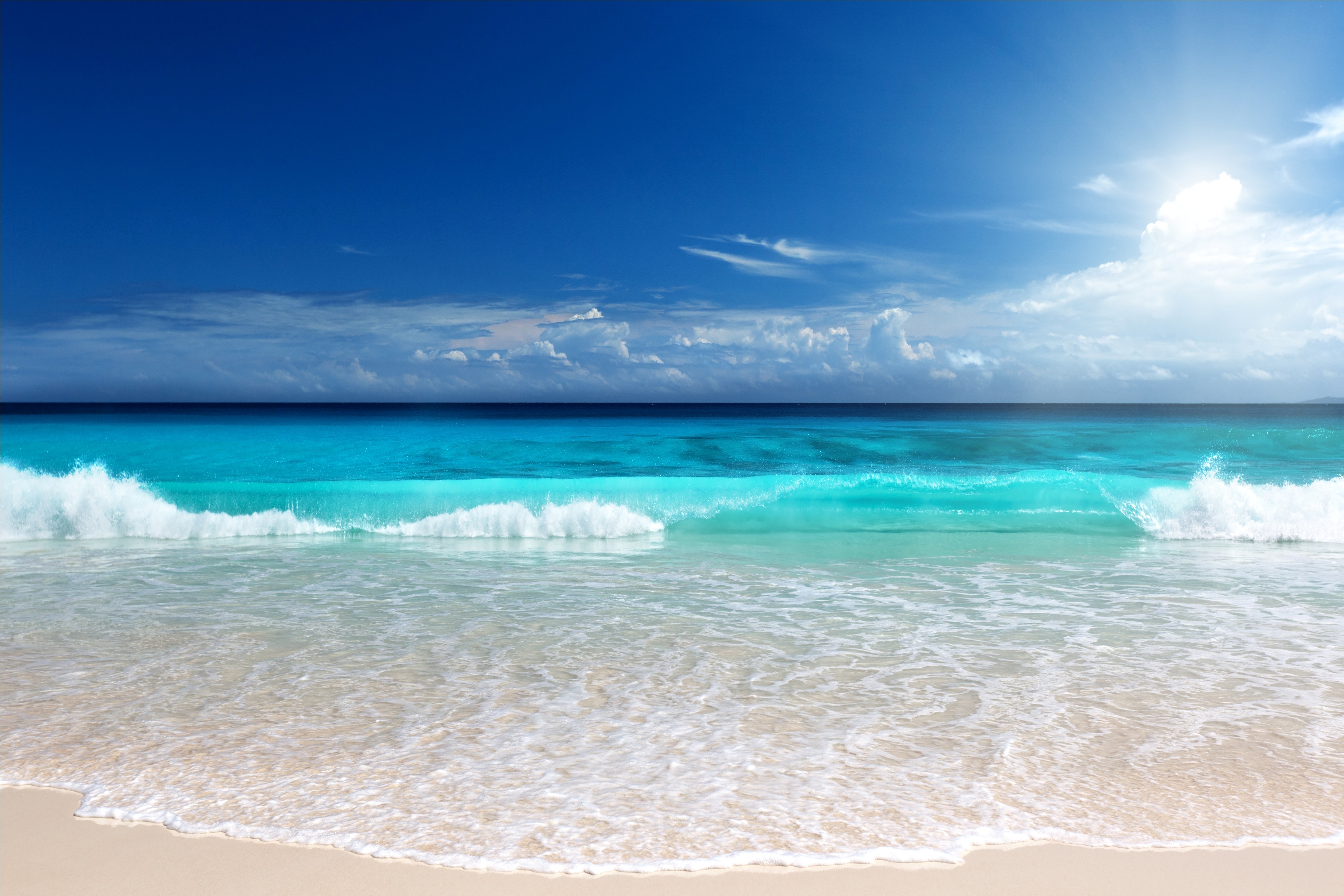 Turquoise ocean waves crash on a sandy beach under a blue sky with clouds and sunlight. HD Wallpaper