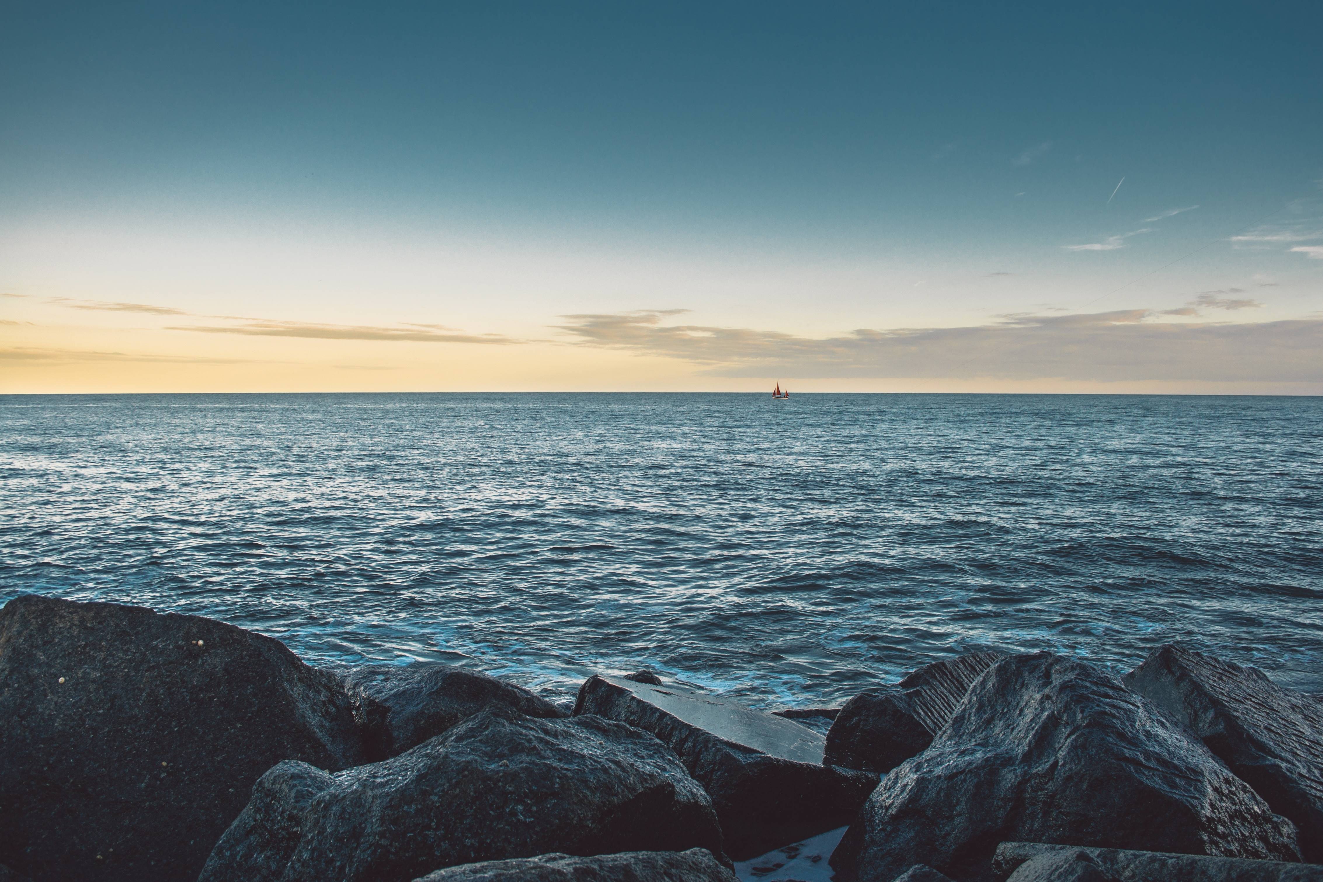 Sailboat on the horizon over the sea with rocks in the foreground HD Wallpaper