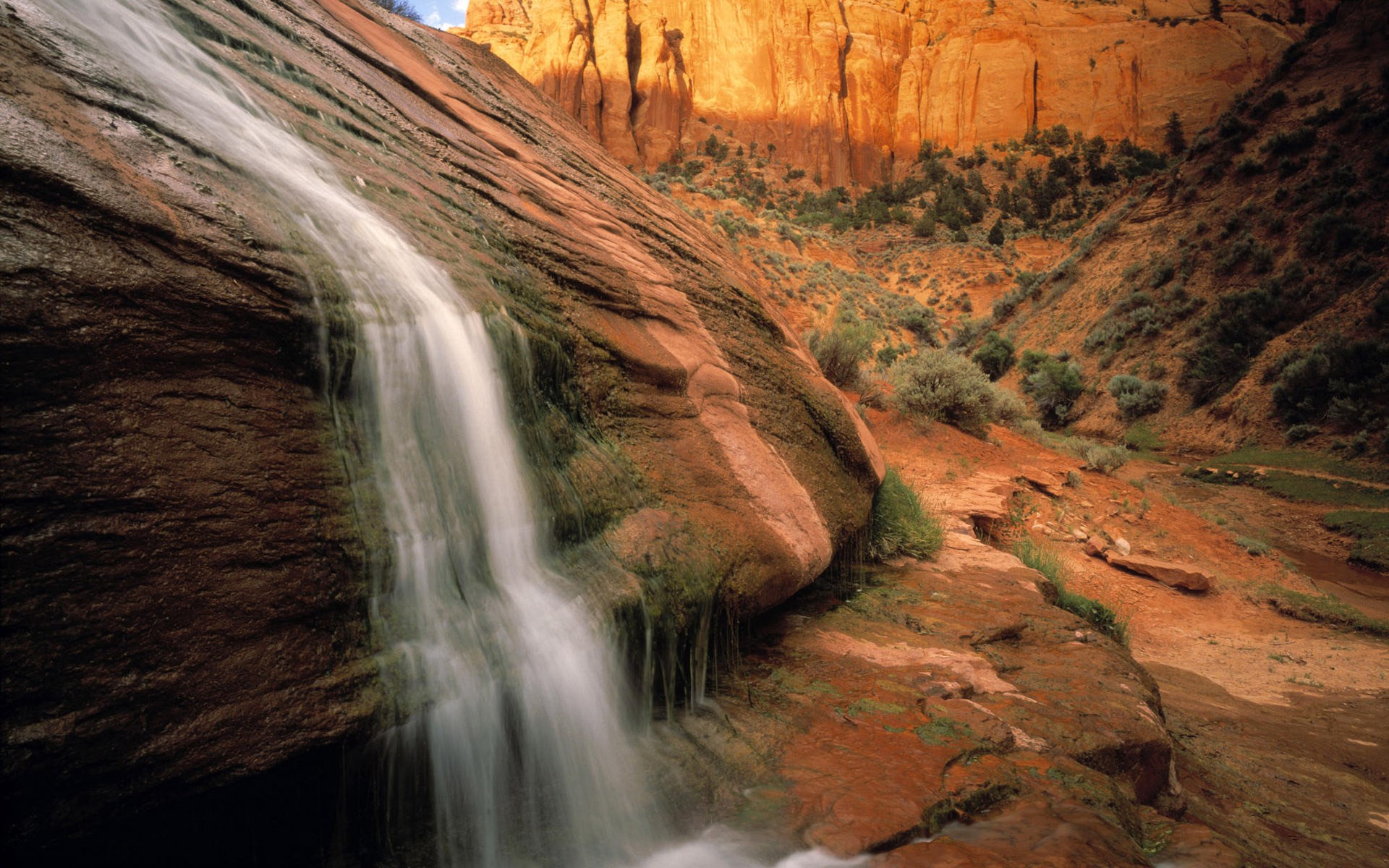Waterfall cascading down a sandstone cliff in a desert canyon HD Wallpaper