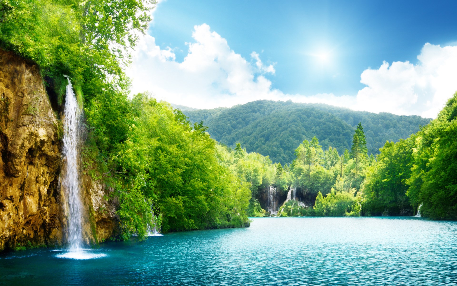 Waterfall cascading into a turquoise lake surrounded by green forest HD Wallpaper
