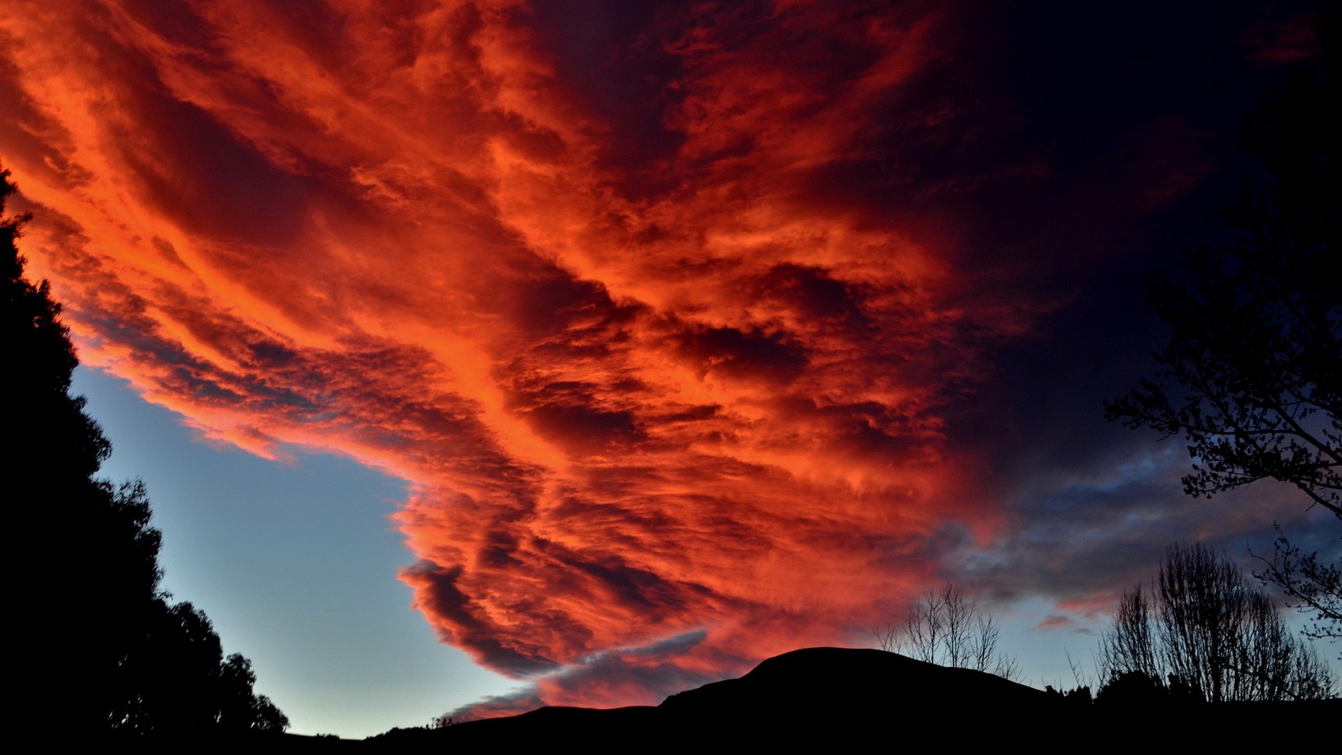 Fiery sunset clouds over silhouetted hills and trees HD Wallpaper