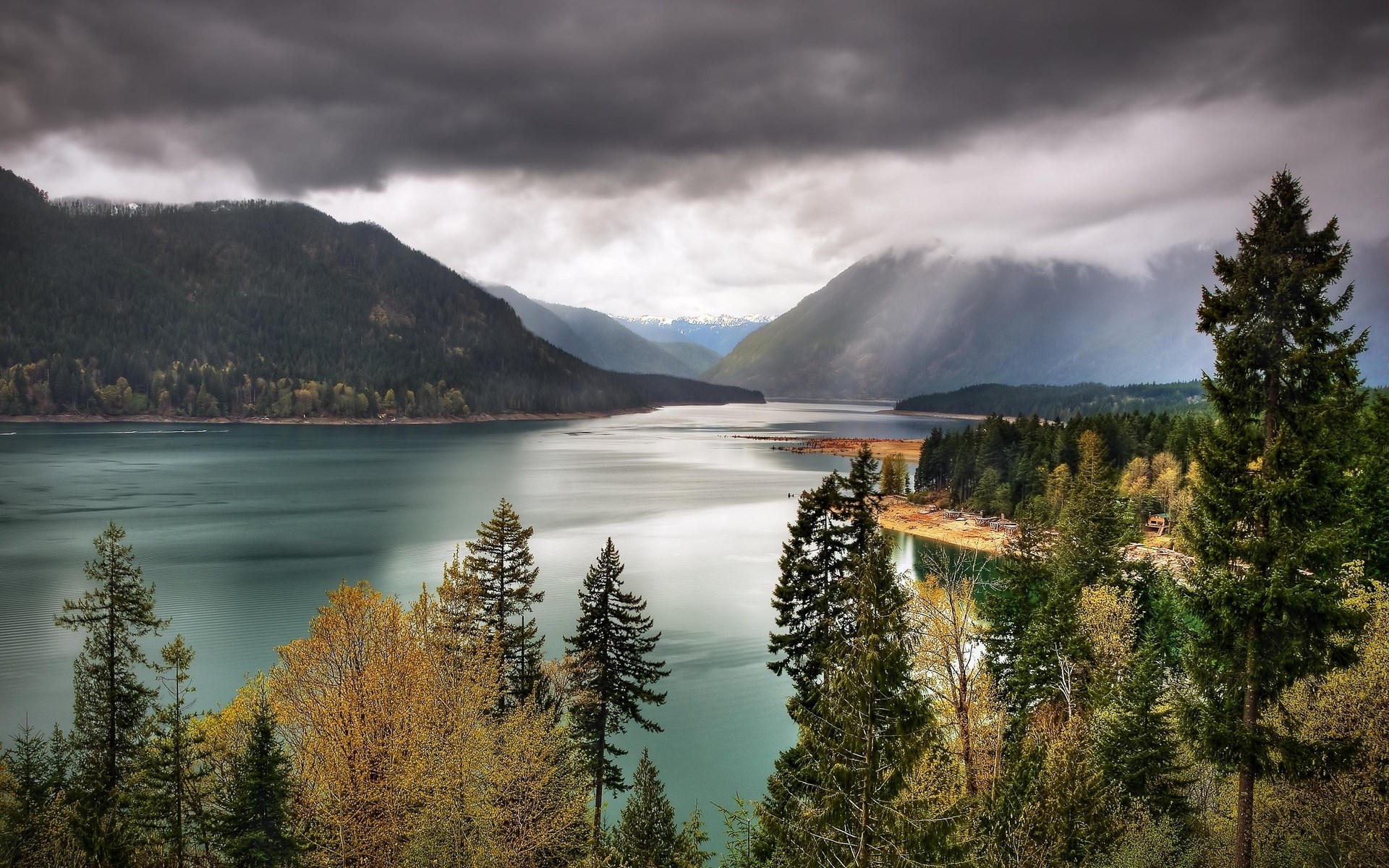 Lake surrounded by mountains and evergreen trees under cloudy sky HD Wallpaper