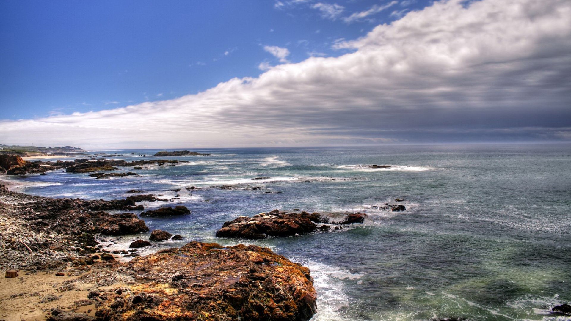 Rocky coastline with waves crashing under a cloudy sky HD Wallpaper