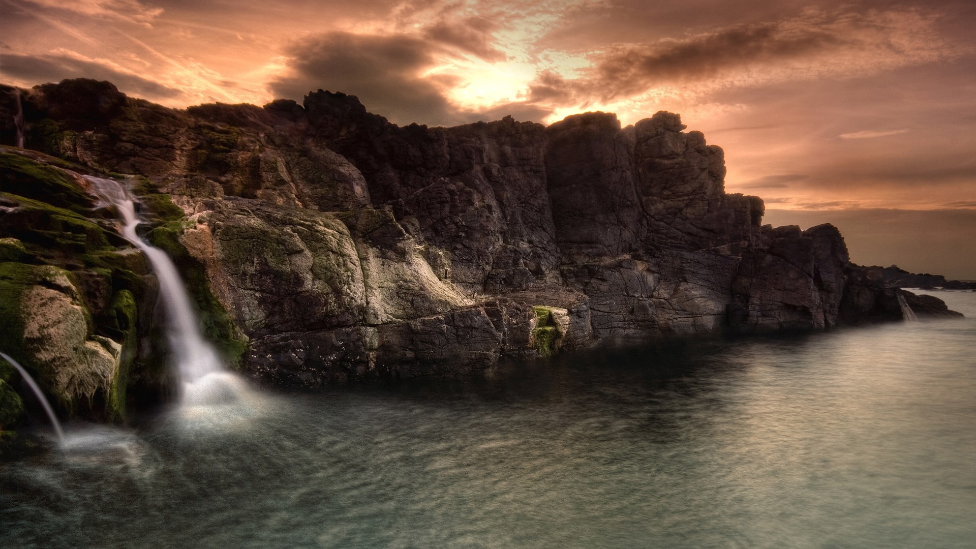 Waterfall cascading over rocky cliffs into the sea at sunset HD Wallpaper