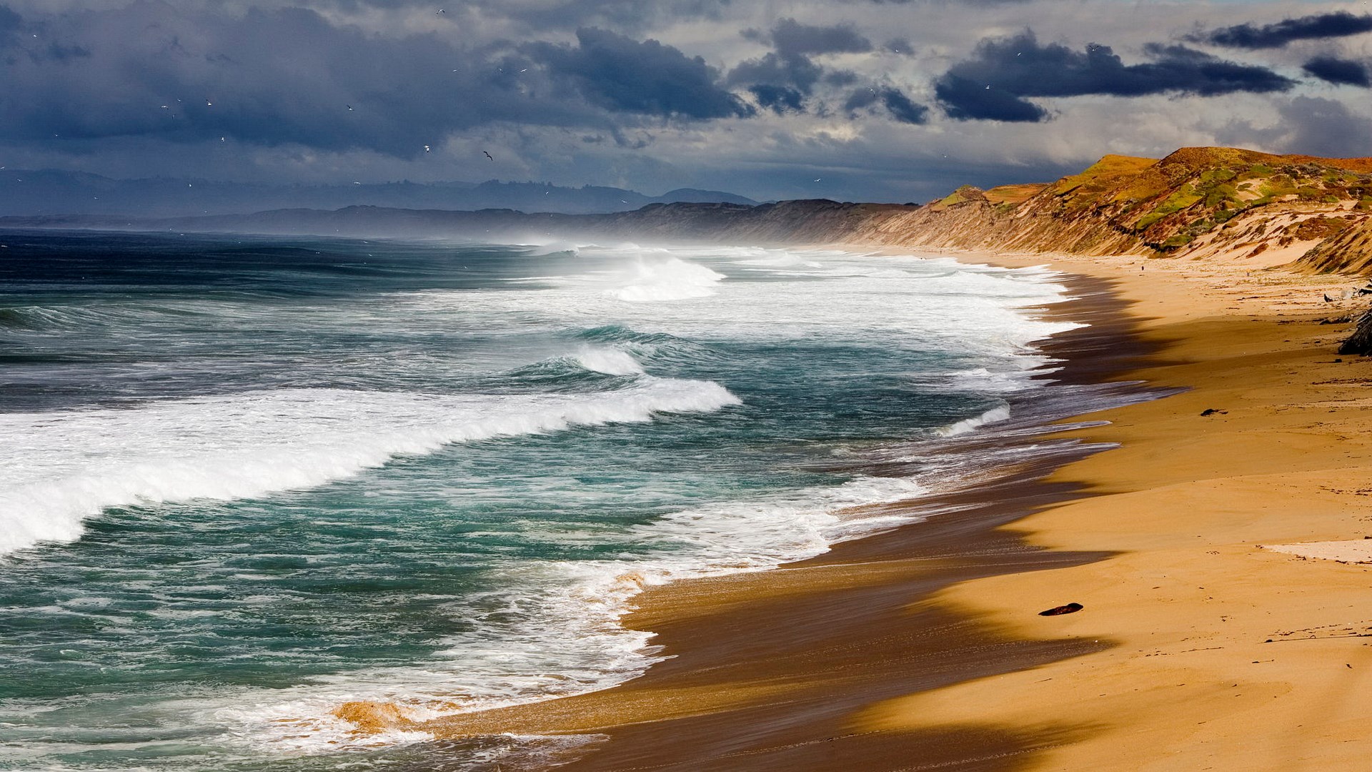 Waves crash on a sandy beach with rolling hills HD Wallpaper
